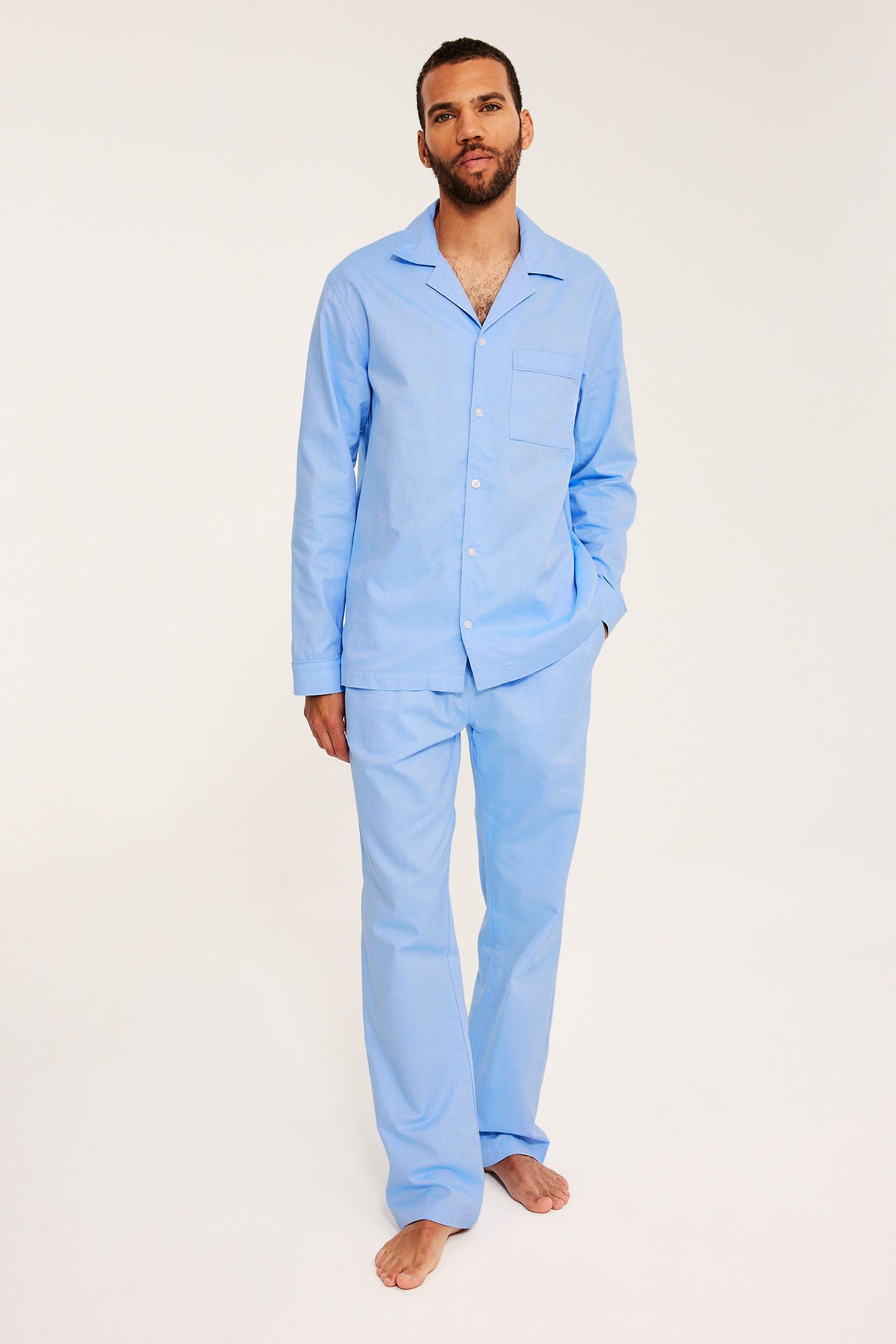 Framifrån, en man i en ljusblå Oxford bomullspyjamasskjorta med knappar och matchande byxor.