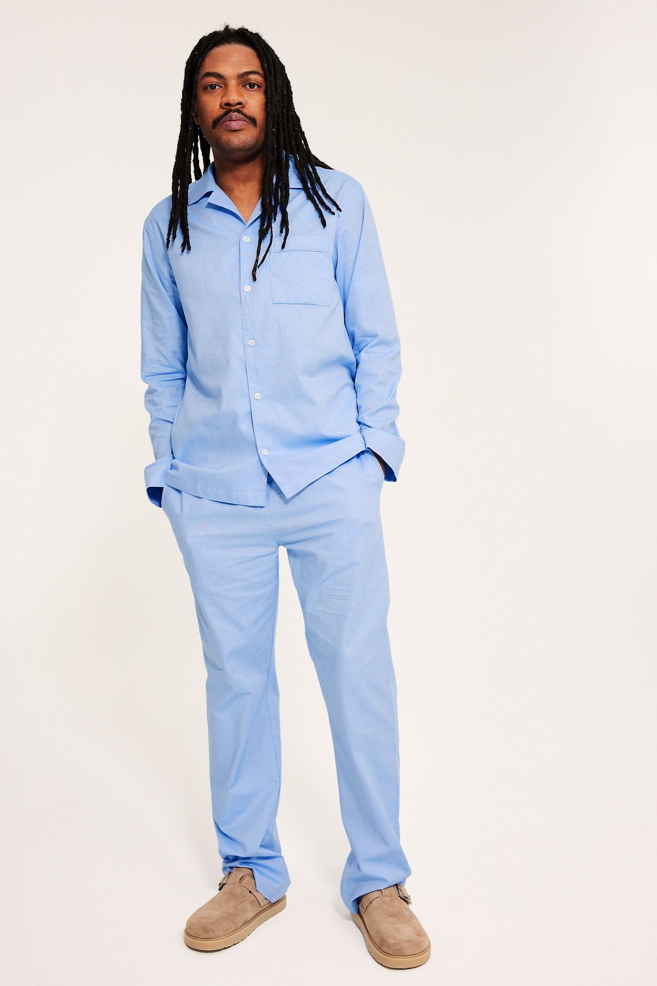 Front: Mann i lyseblå bomulls-Oxford-pyjamas med knappeskjorte, brystlomme og bukser med elastisk midje. Brune tøfler.