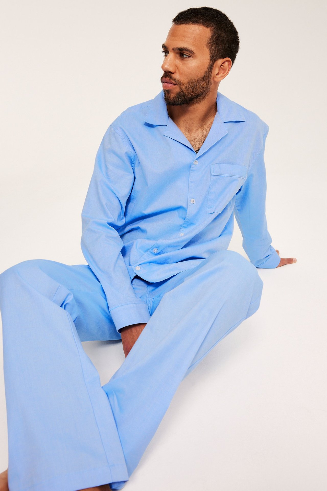 Edestäpäin istuva mies yllään vaaleansininen Oxford-puuvillapyjama napeilla ja rintataskulla.