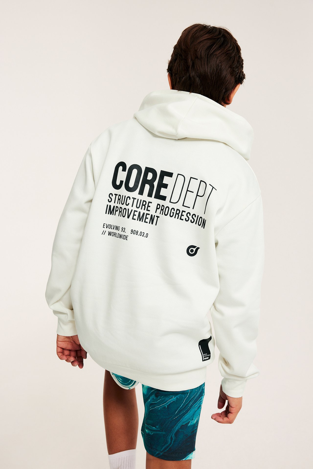 Widok z tyłu na dziecko w białej bluzie z kapturem z czarnym nadrukiem i turkusowych szortach.