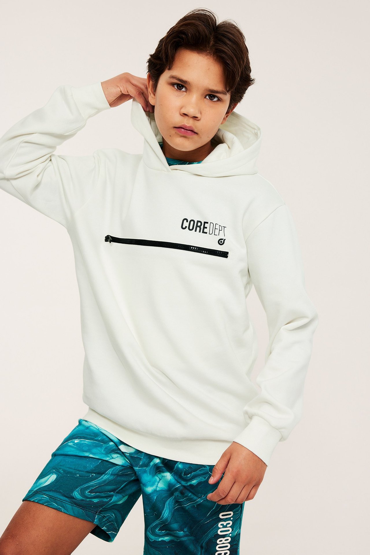 Widok z przodu: Dziecko w białej bluzie z kapturem z zamkiem, nadrukiem "COREDEPT" i turkusowymi szortami.