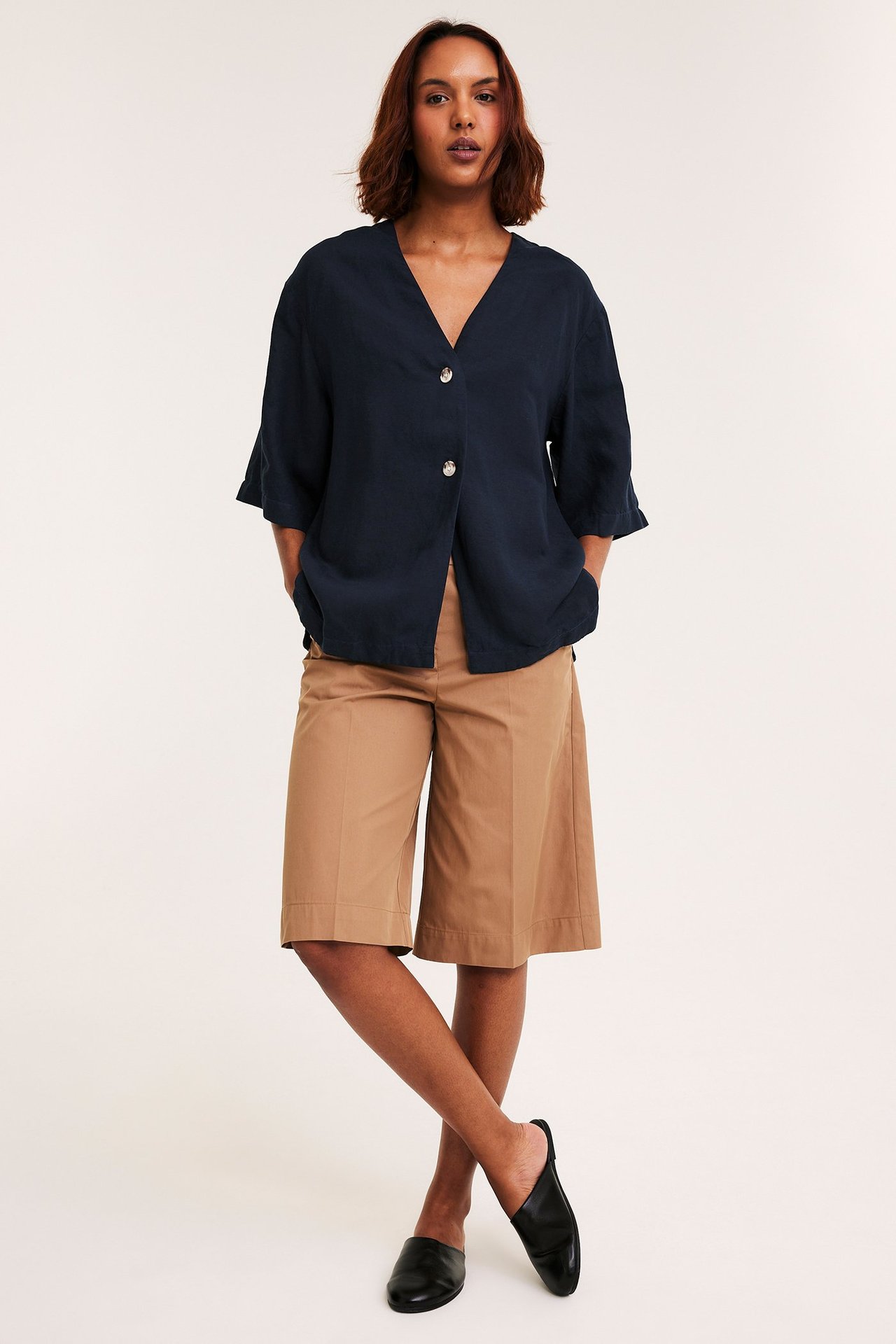 Nainen edestä, yllään tummansininen pellavasekoite lyhythihainen paita, beiget leveät shortsit ja mustat muulit.