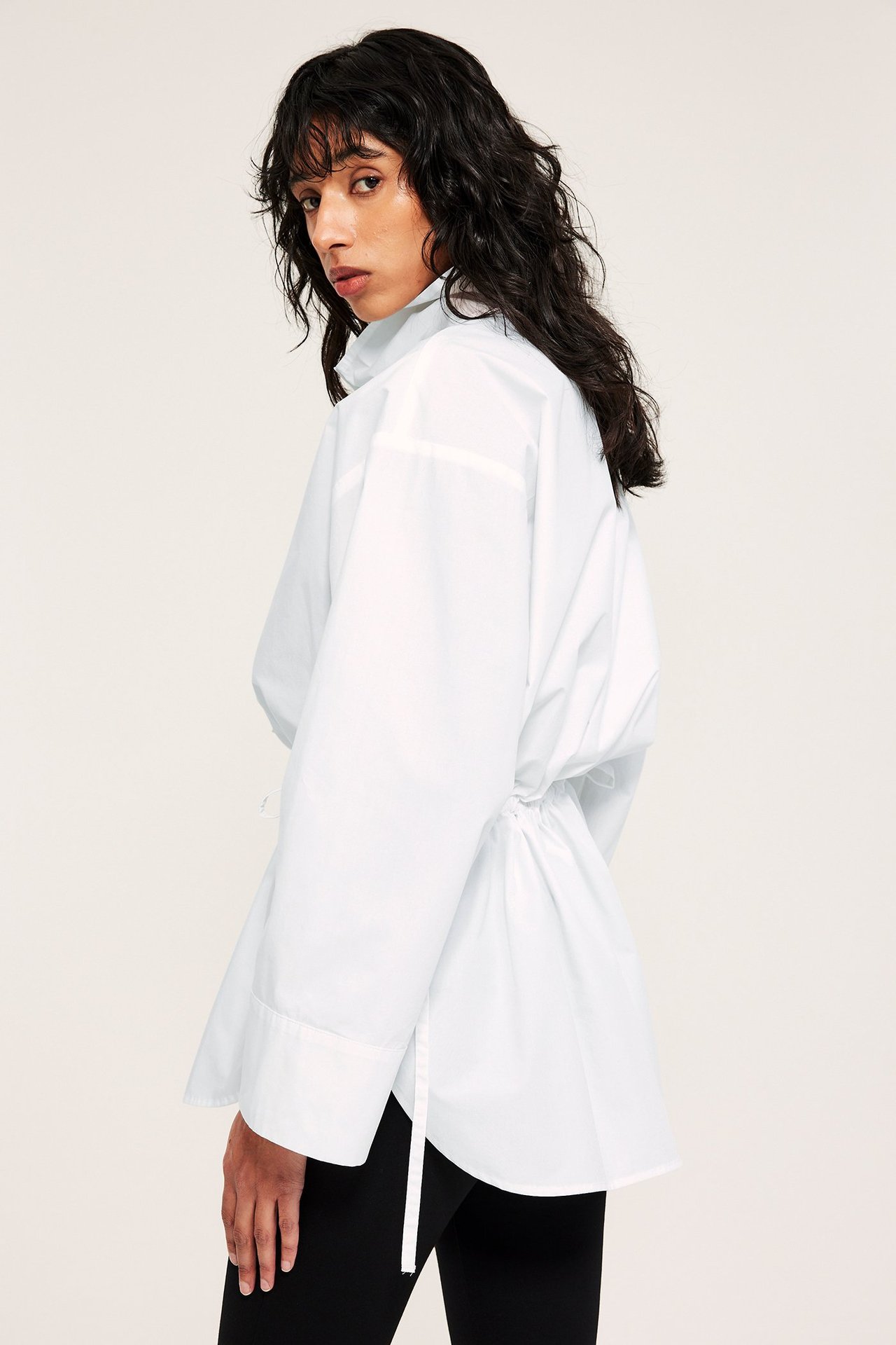 Sidovy: kvinna i vit poplinskjorta med ståkrage och dragsko i midjan, med svarta byxor.