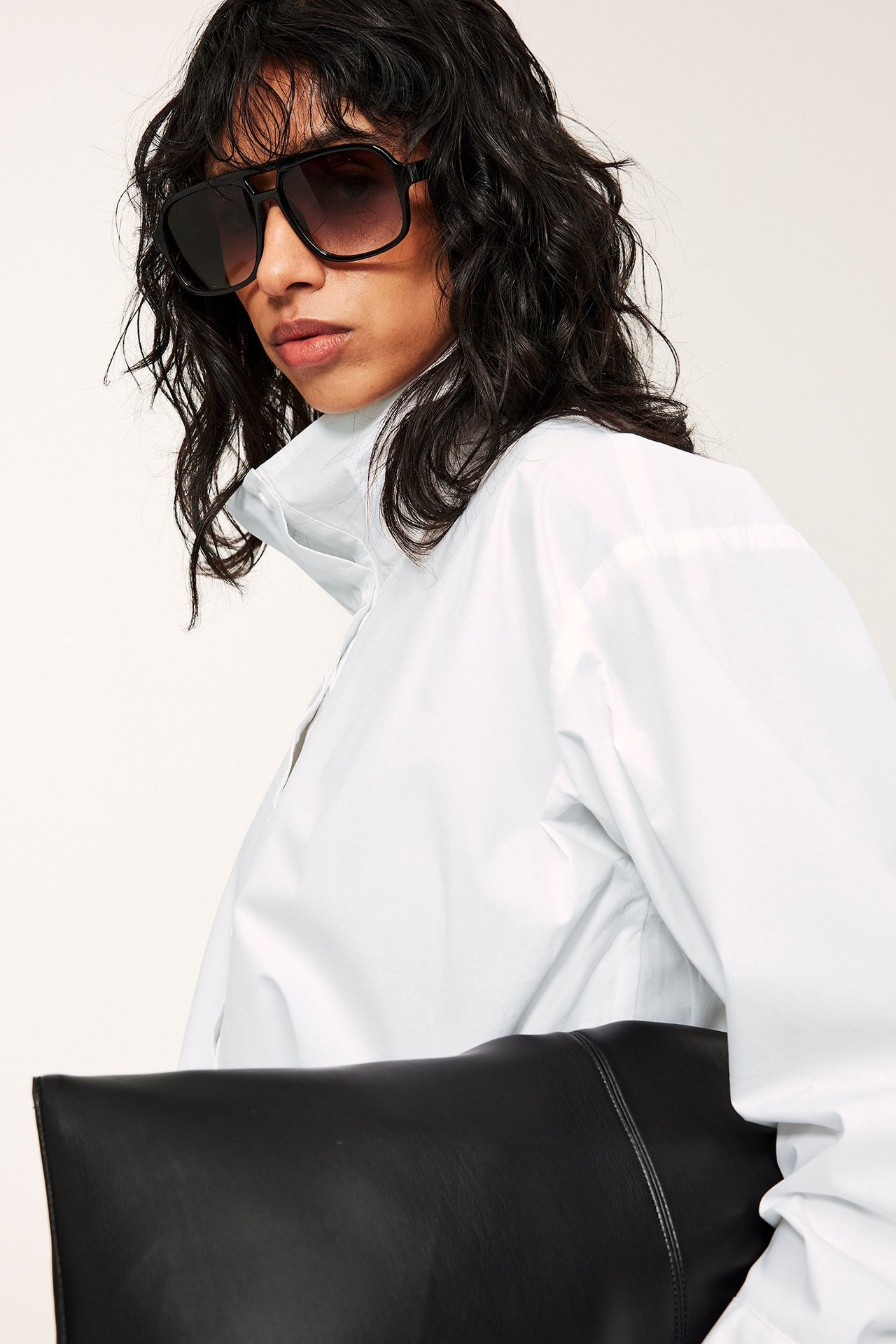 Närbild av en kvinna i en vit poplinskjorta med ståkrage, hållande en svart clutch och solglasögon.