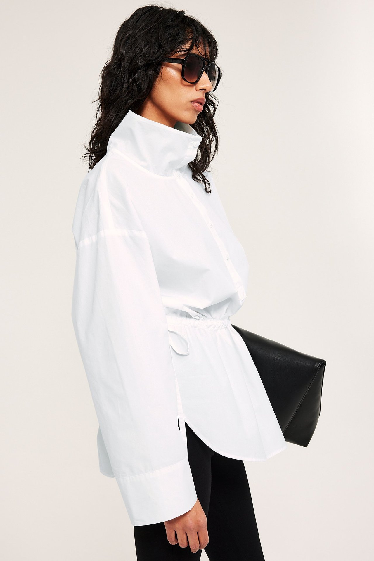 Sidovy av en kvinna i vit poplinskjorta med ståkrage, dragsko i midjan, svarta solglasögon och svart clutch.