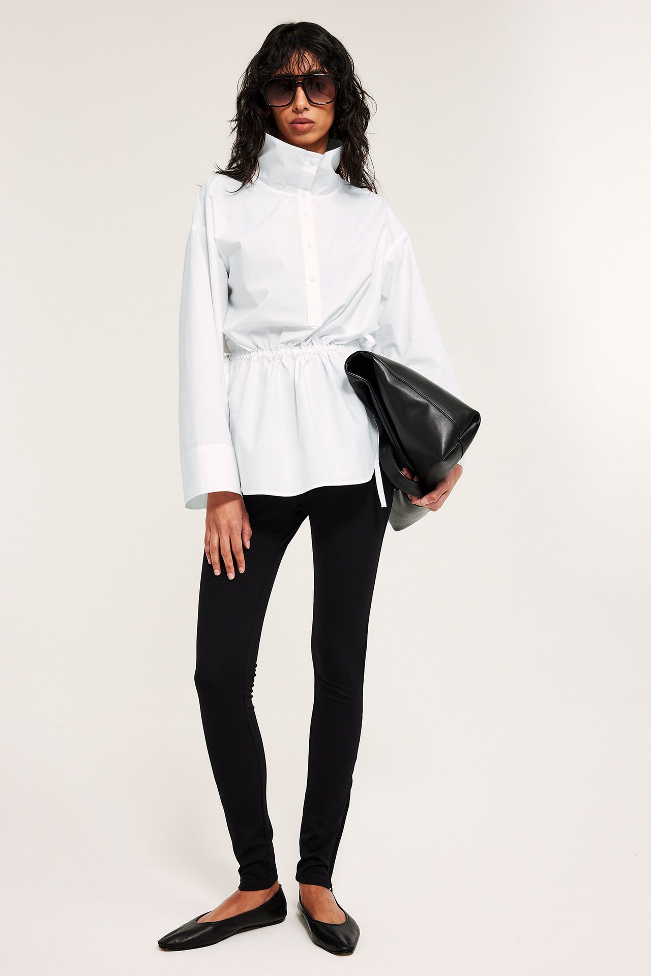 Framifrån: kvinna i vit poplinskjorta med dragsko i midjan, svarta leggings och håller en svart handväska.