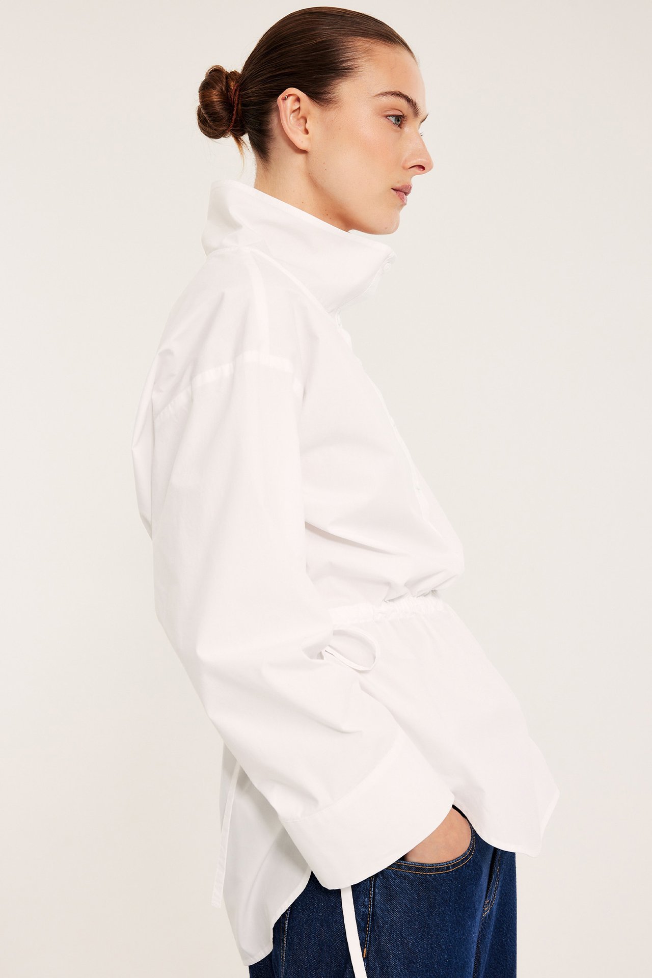 Widok z boku kobiety w białej koszuli popelinowej ze stójką, ściągaczem w talii i ciemnoniebieskich dżinsach.
