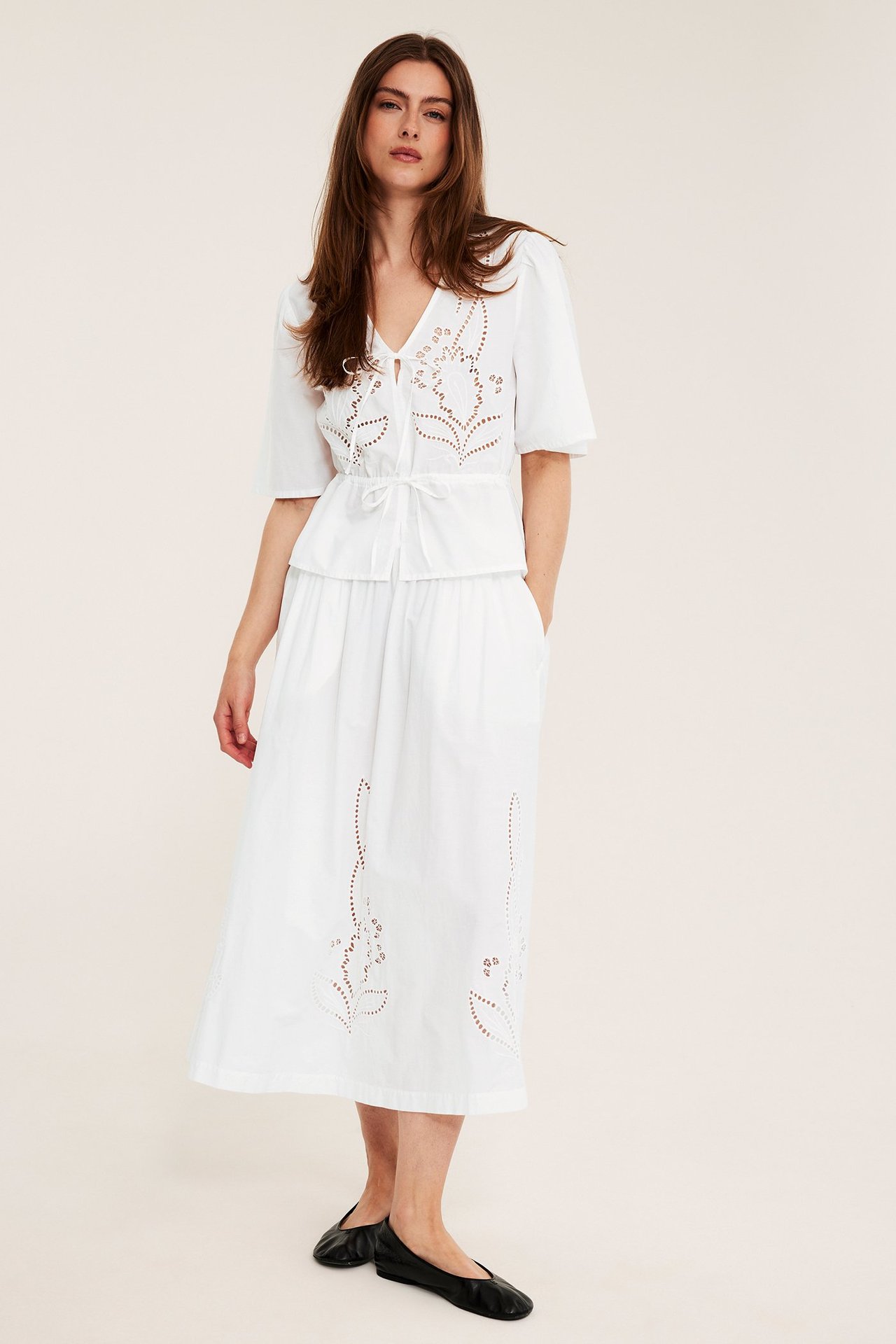 Framsidan av en kvinna i vit broderad bomullspoplinblus, matchande kjol och svarta ballerinaskor.