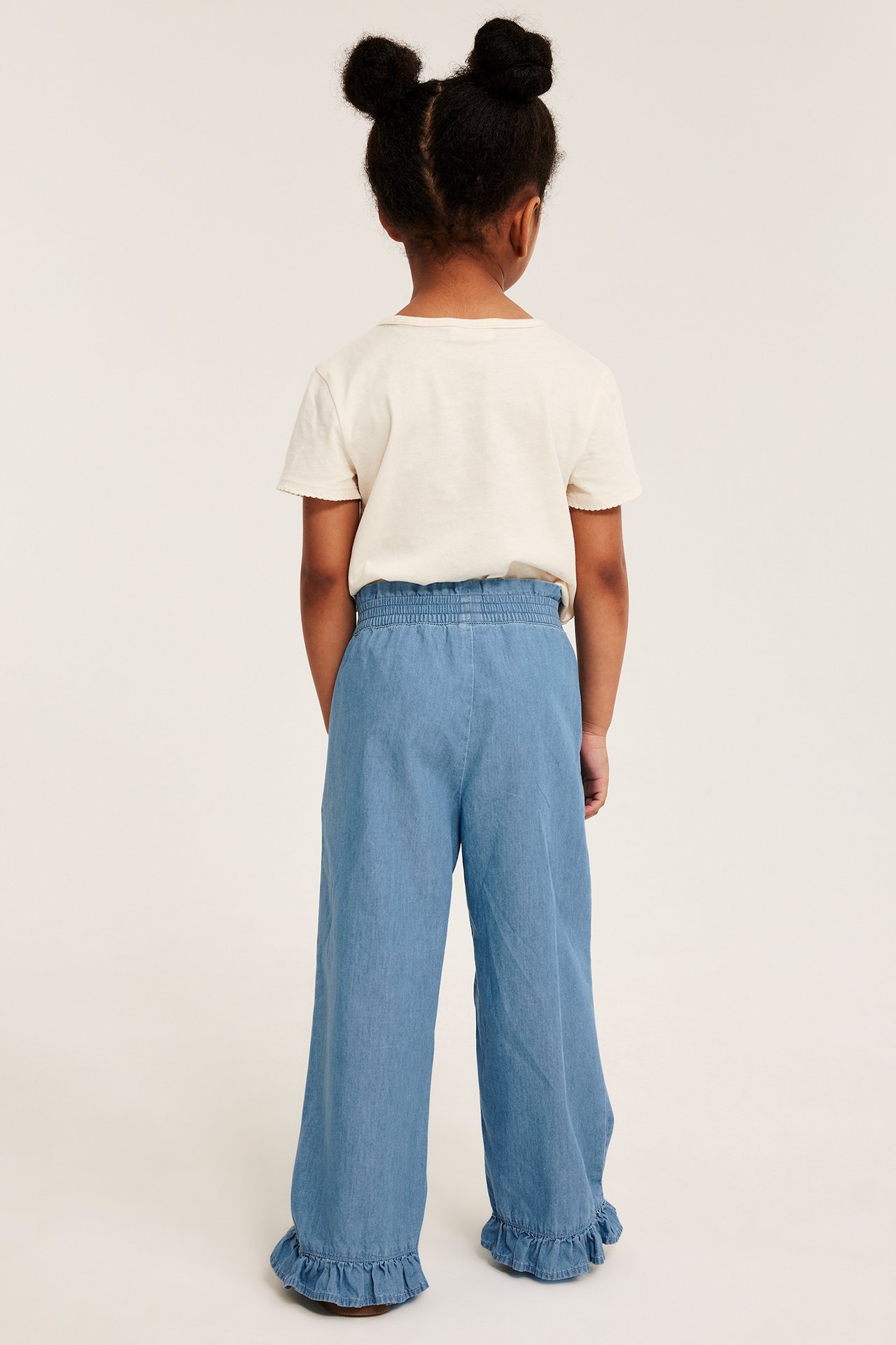 Widok z tyłu dziecka w jasnoniebieskich szerokich dżinsowych spodniach z falbankami i jasnym T-shircie.