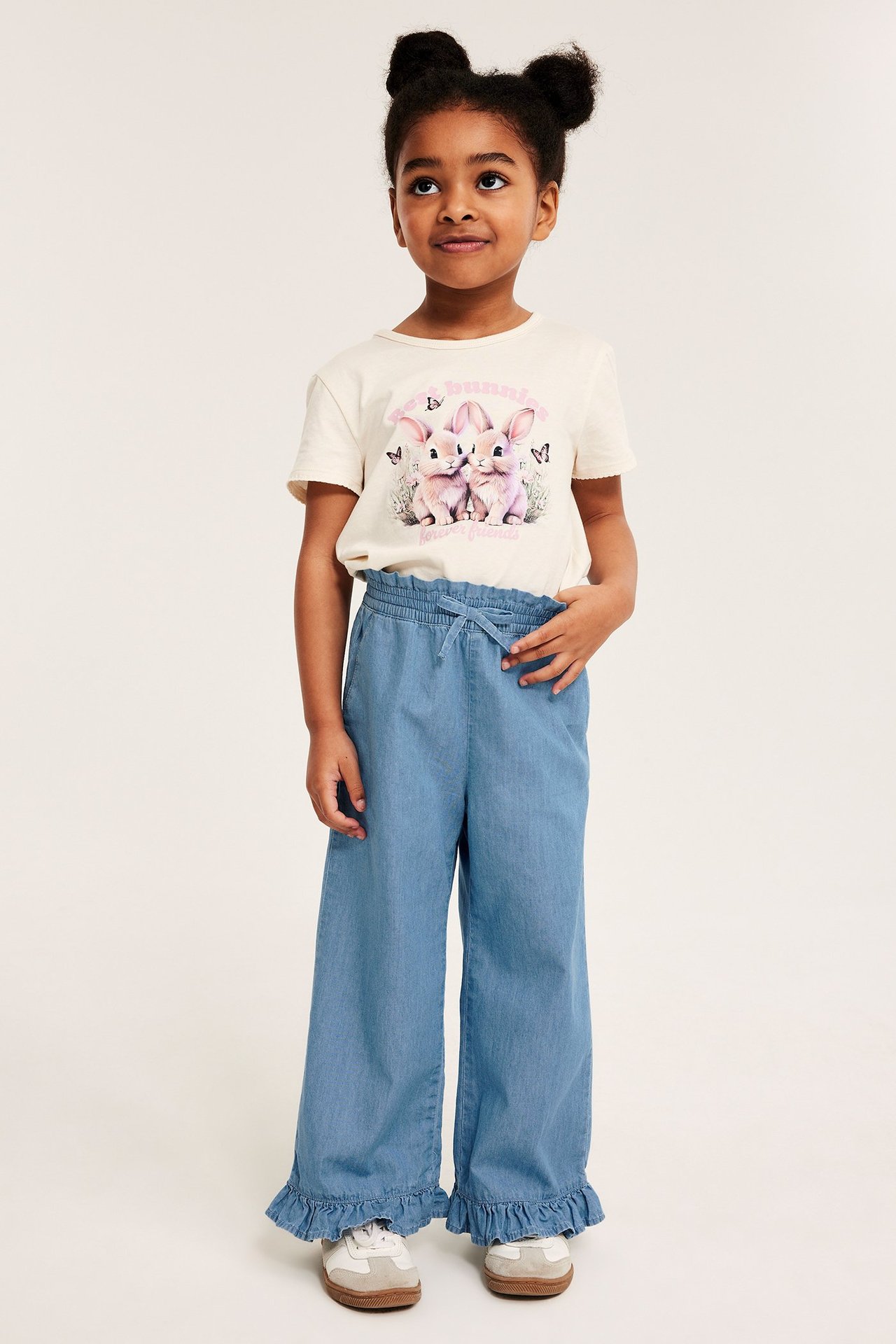 Frontvy: Kid i ljusblå vida denimbyxor med volang nertill och en cremevit t-shirt med kanintryck.