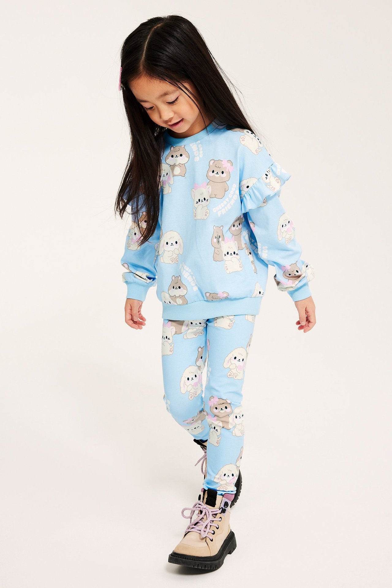 Edestä näkymä tytöstä vaaleansinisessä eläinkuvioidussa puuvillapaidassa röyhelöillä, yhteensopivissa leggingseissä ja beigenvärisissä saappaissa.