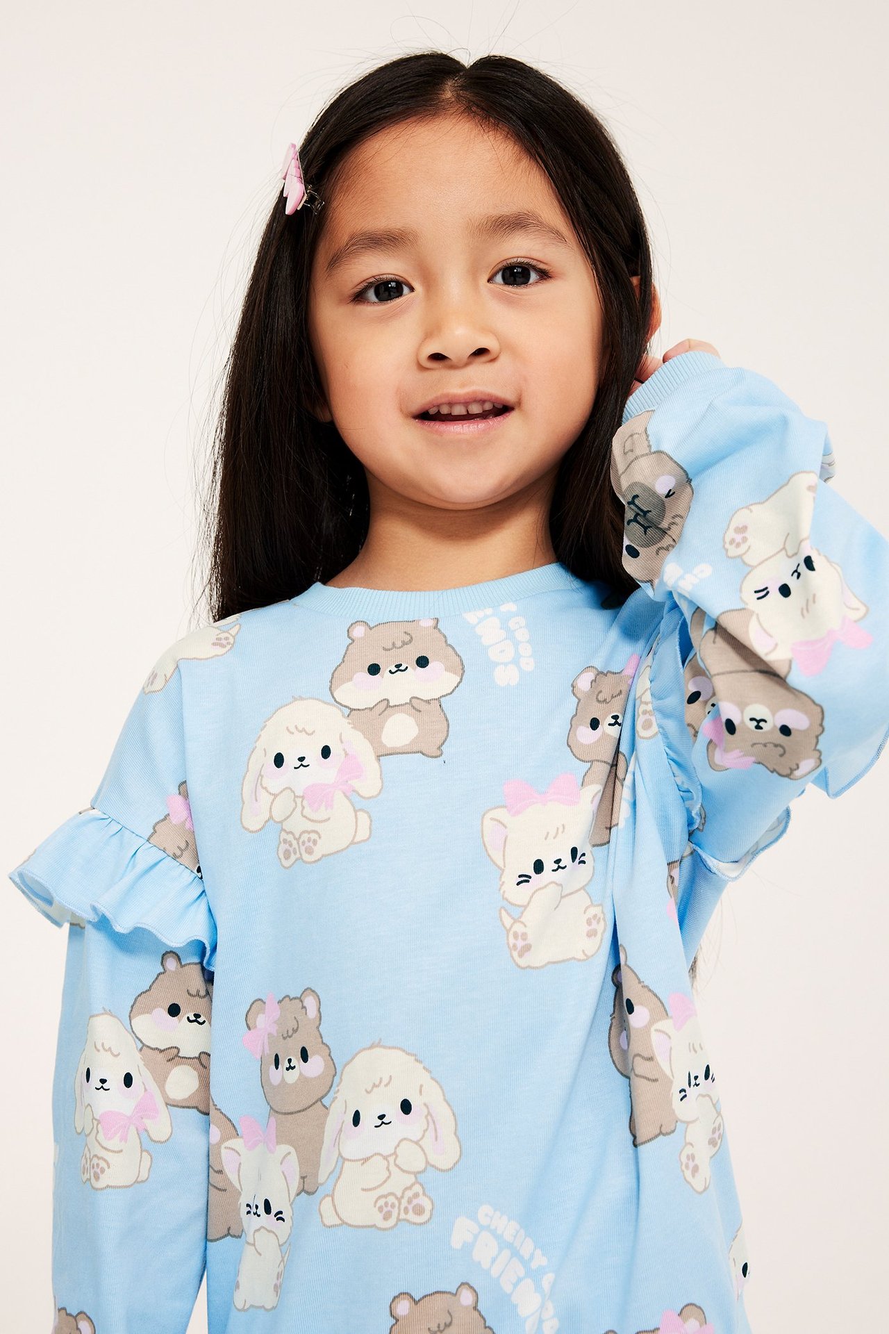 Widok z przodu: Dziecko w jasnoniebieskiej bluzie bawełnianej z nadrukiem zwierzęcym, falbankami i różową spinką.