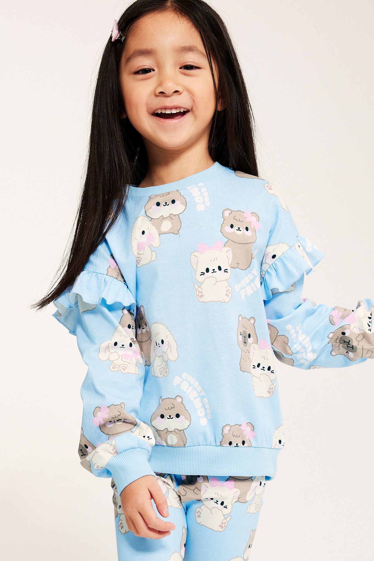 Z przodu: Uśmiechnięte dziecko w jasnoniebieskiej bluzie bawełnianej w zwierzęcy wzór, z falbankami na rękawach i legginsach.