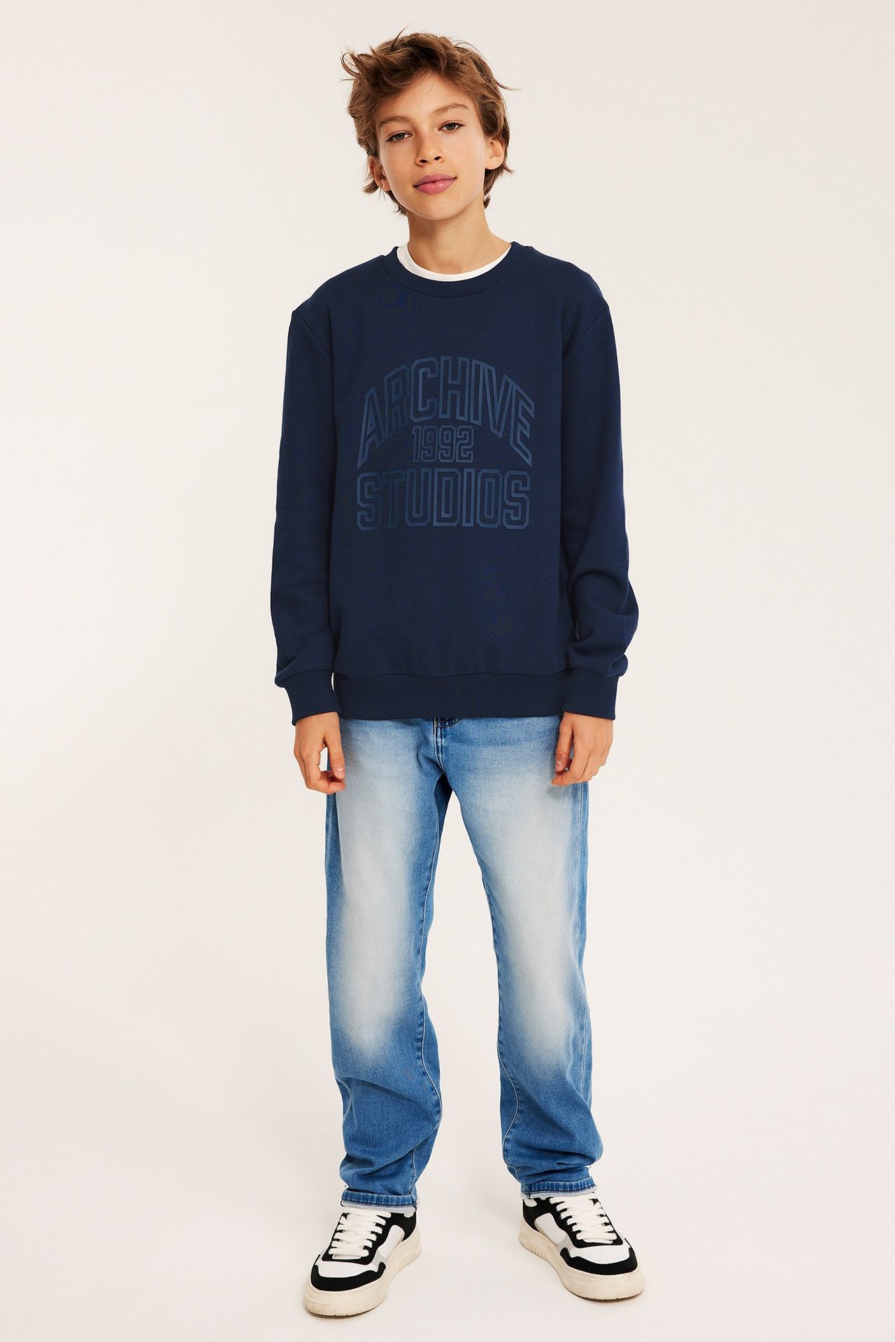 Poika yllään tummansininen painettu collegepaita, valkoinen t-paita, vaaleansiniset farkut ja mustavalkoiset tennarit.