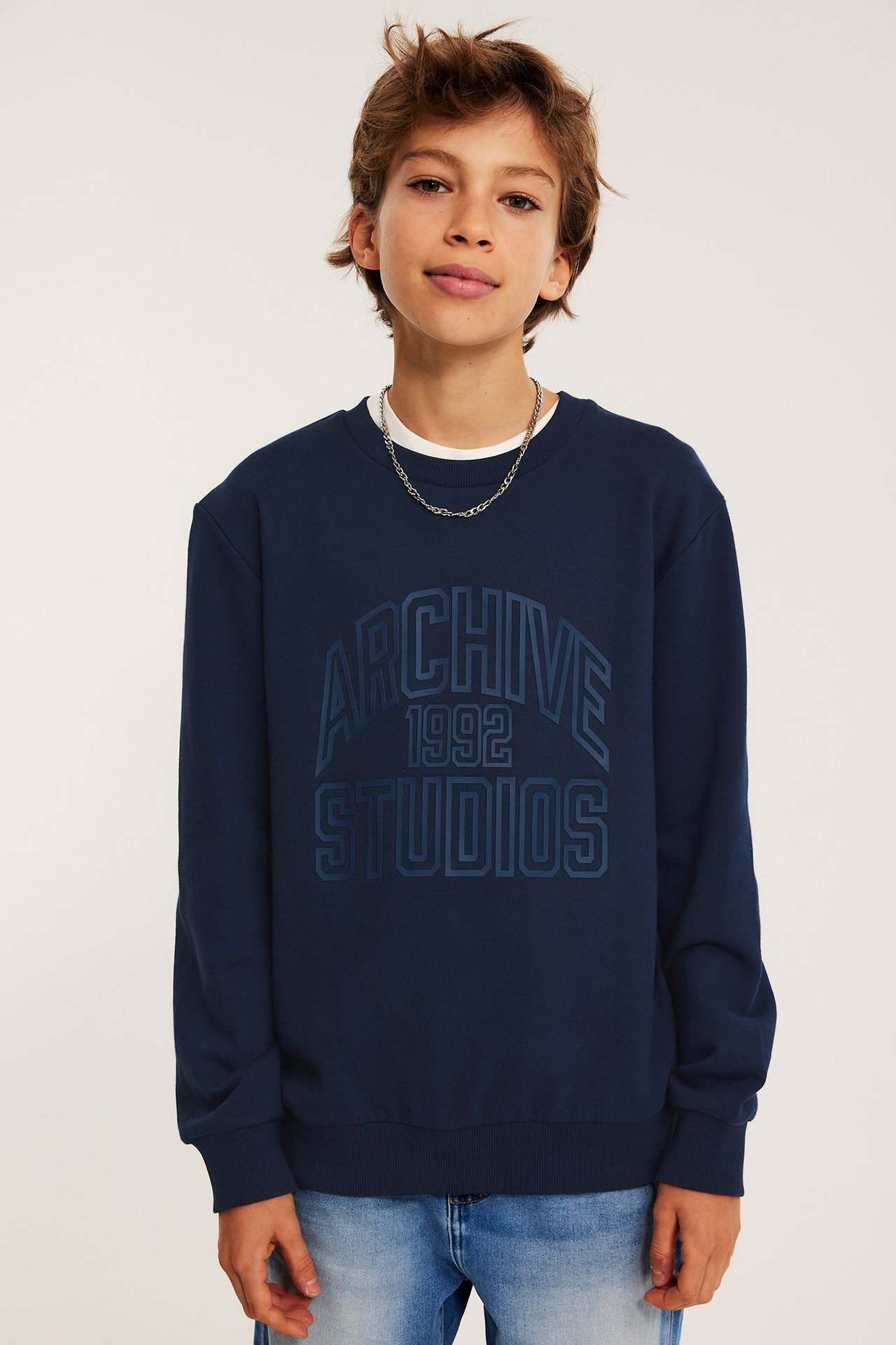 Widok z przodu: Dziecko nosi granatową bawełnianą bluzę z nadrukiem, białą koszulkę, niebieskie dżinsy, srebrny naszyjnik.