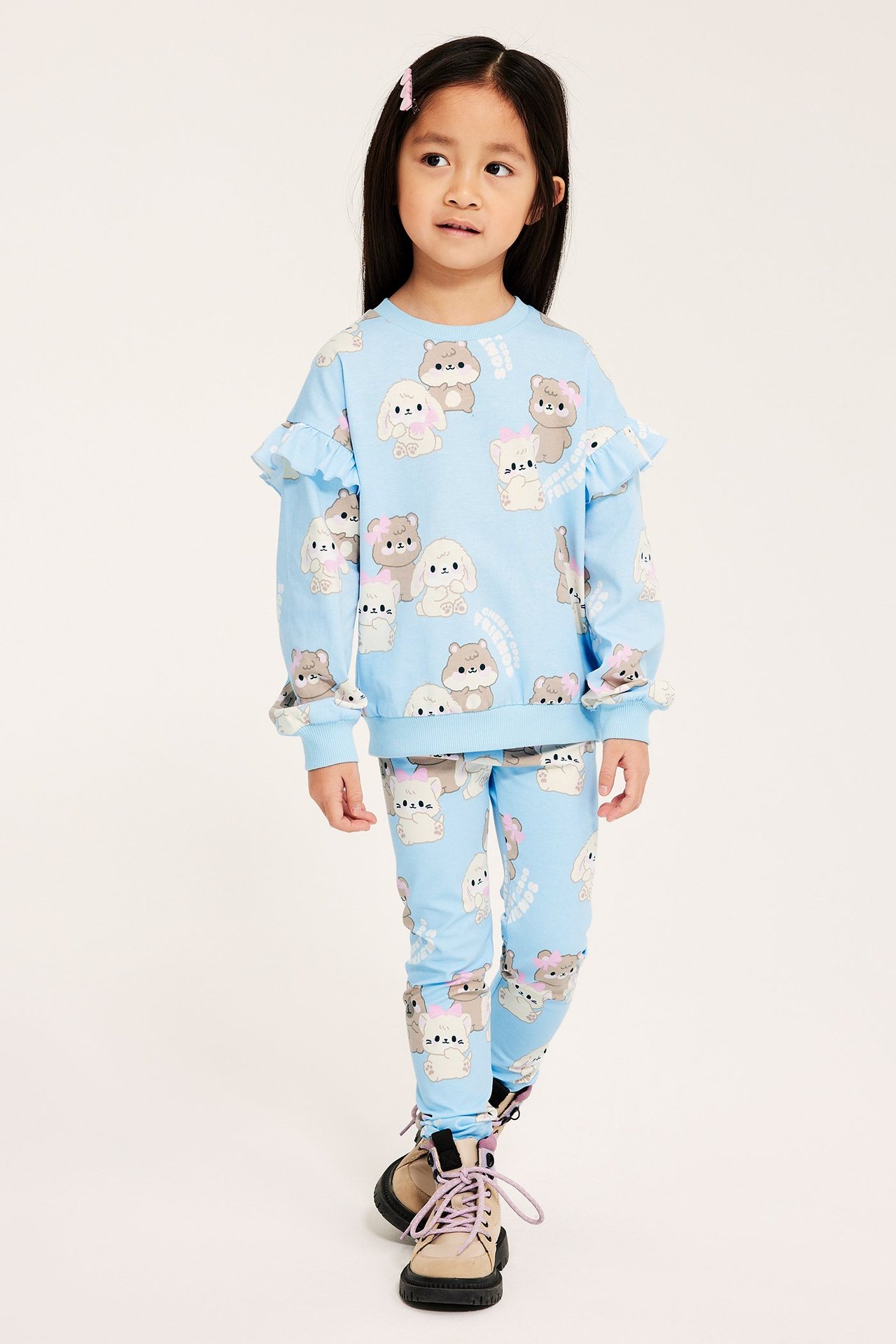 Widok z przodu: dziecko w jasnoniebieskich legginsach we wzory, pasującej bluzce z falbankami i beżowych butach.