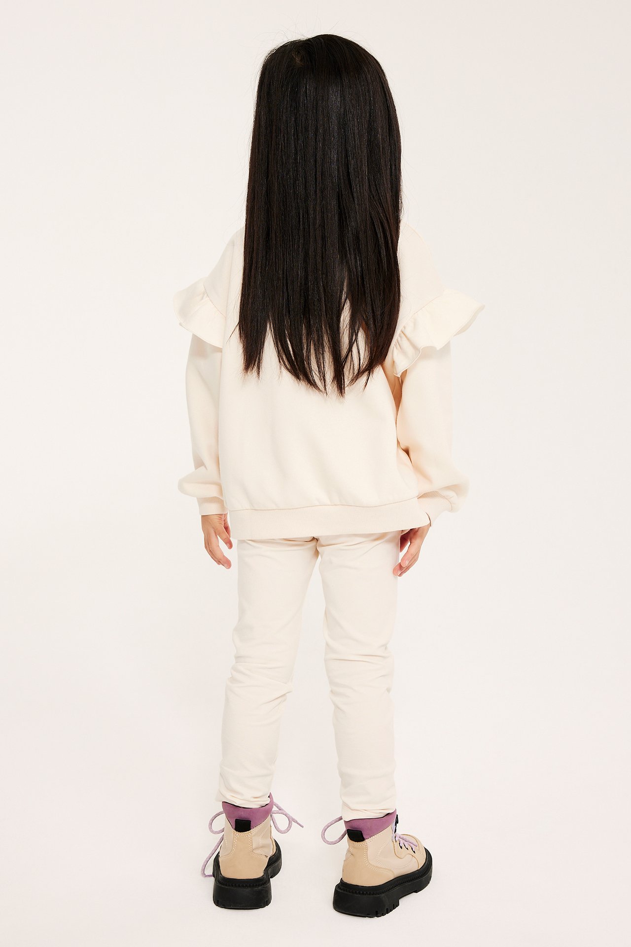 Takakuva lapsesta: luonnonvalkoiset leggingsit, röyhelöpusero ja beiget kengät violetilla nauhoilla.