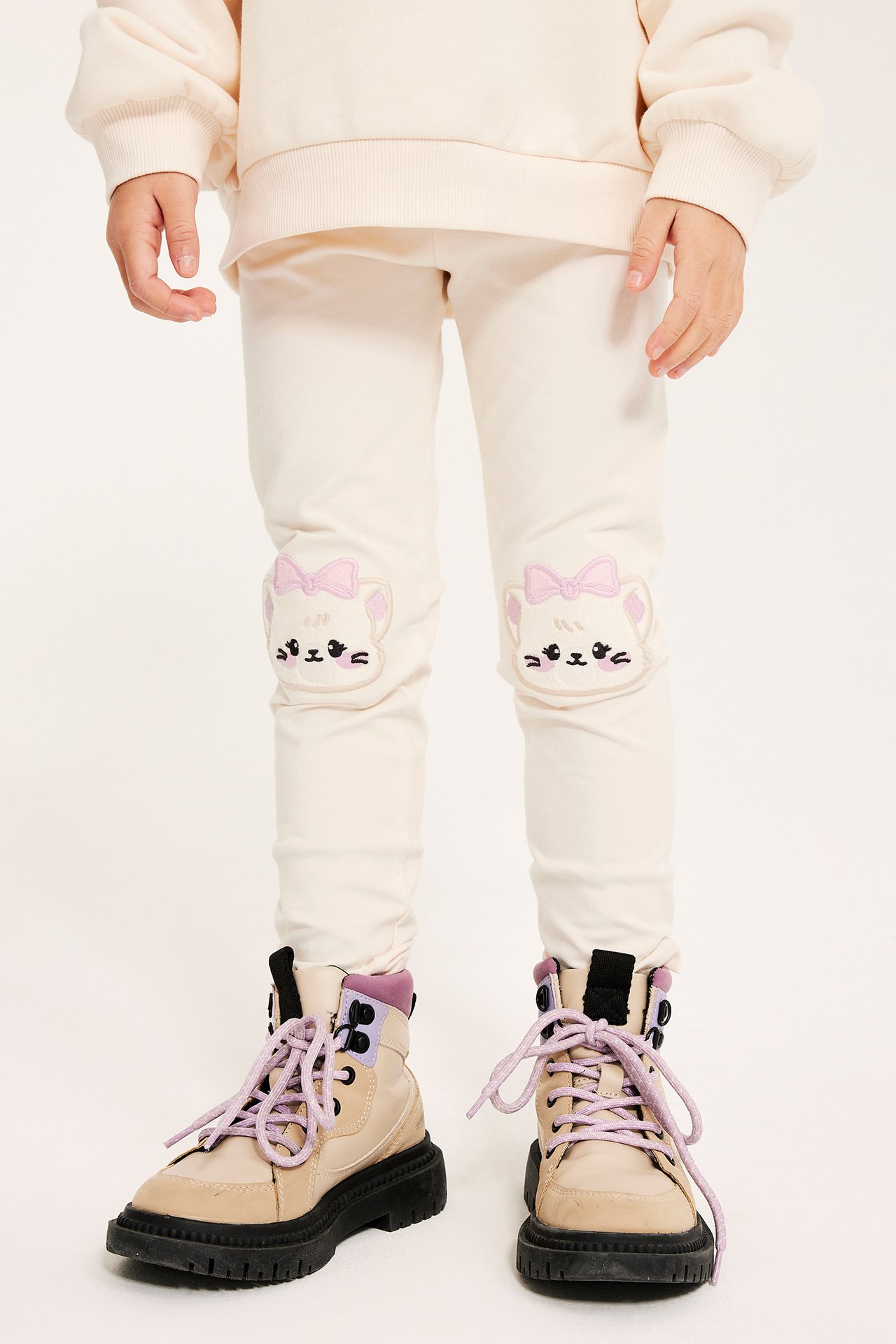 Framifrån: Barn bär benvita leggings i bomull med kawaii-kattapplikationer på knäna, en benvit sweatshirt och beiga kängor.