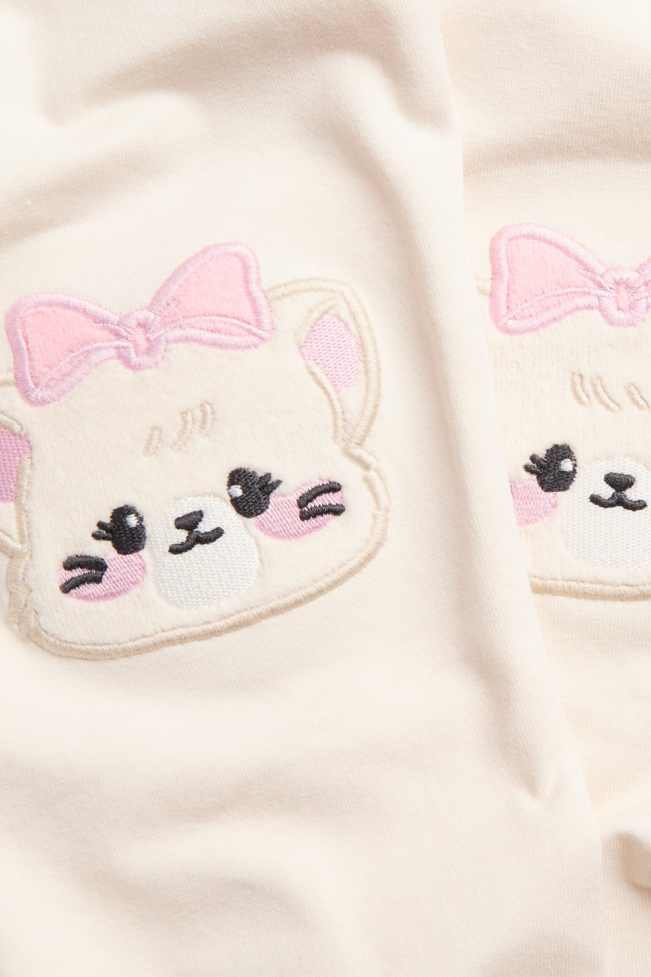 Zbliżenie: Dziecięce legginsy bawełniane w kolorze złamanej bieli z welurowymi aplikacjami głowy misia i różowymi kokardkami na kolanach.