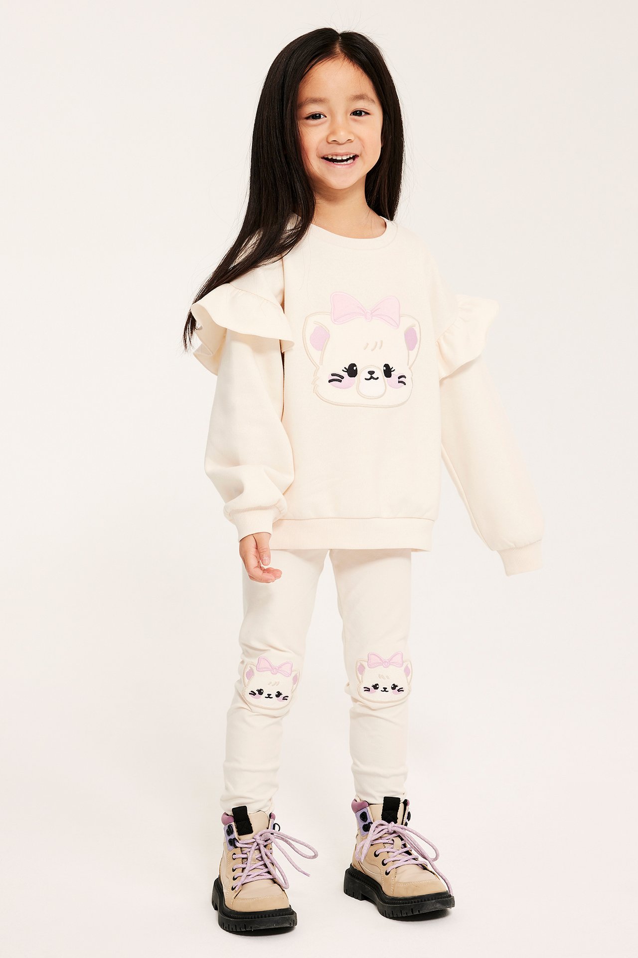 Widok z przodu, dziecko w kremowych legginsach z kotkami na kolanach, pasująca bluza i beżowe buty.