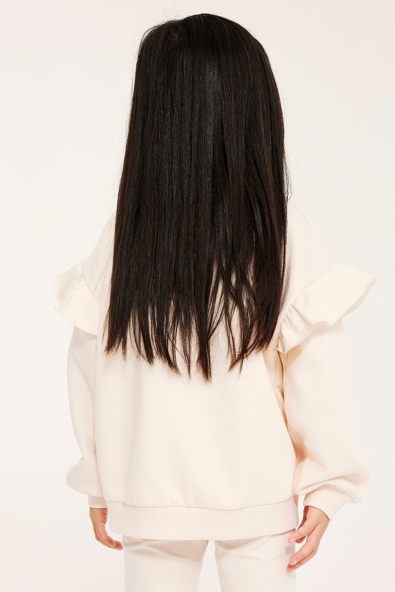 Takakuva lapsesta: luonnonvalkoinen collegepusero röyhelöhihoilla, ribbimudilla ja yhteensopivat housut.