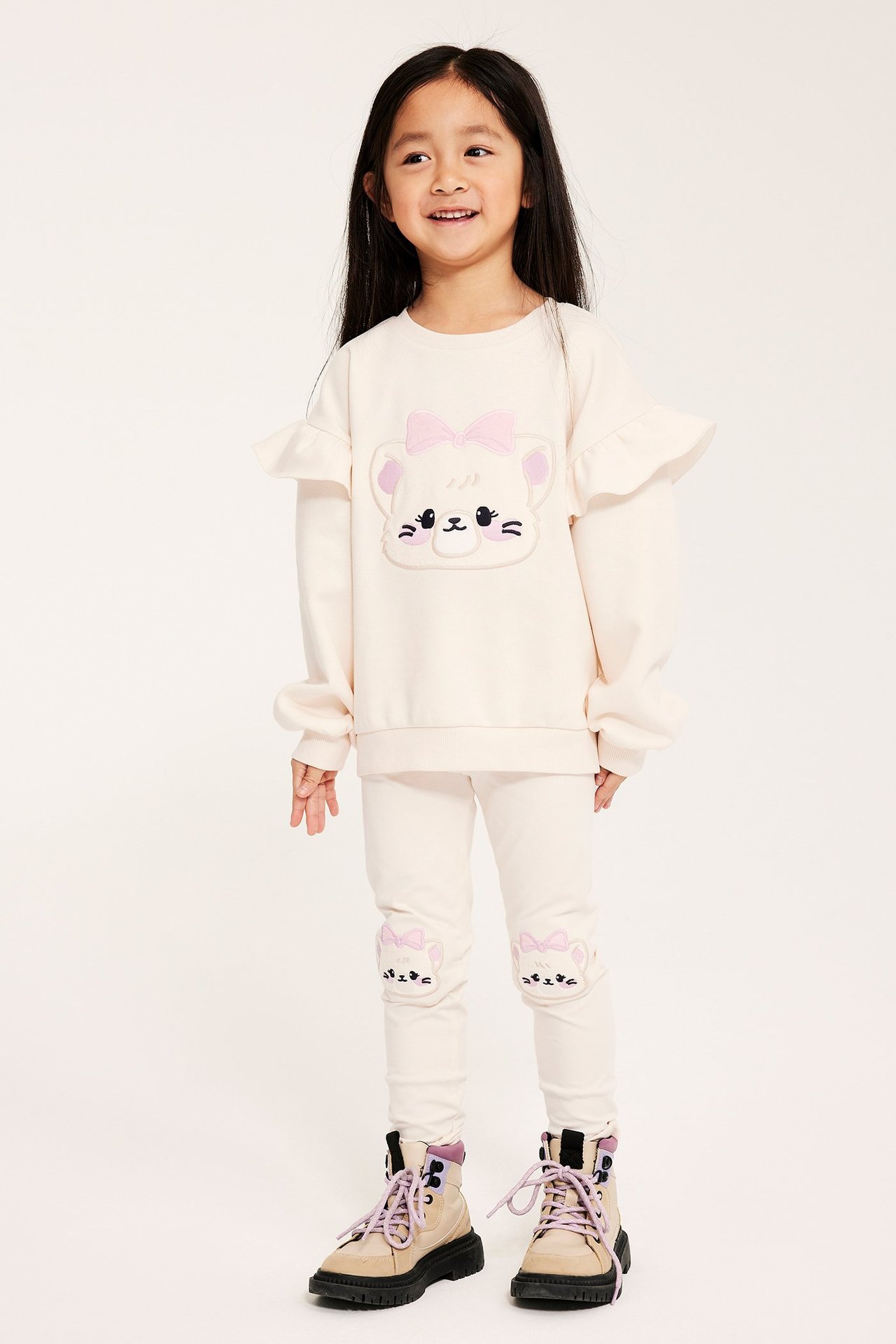 Forfra: Barn har på seg en offwhite sweatshirt med katteapplikasjon, volanger, matchende leggings og støvler.