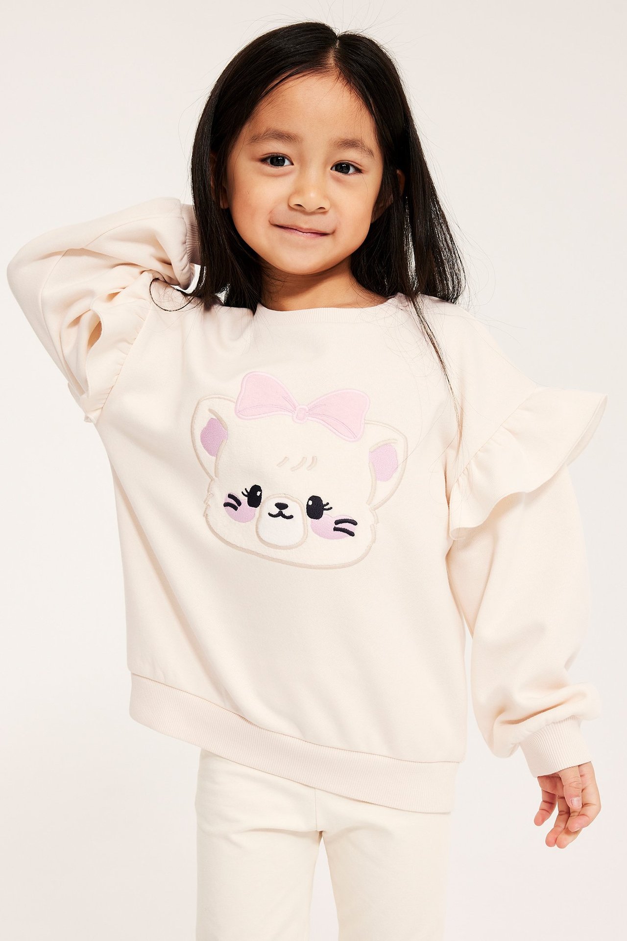 Widok z przodu dziecka w kremowej bluzie z aplikacją kota, falbankami na rękawach i pasujących kremowych spodniach.
