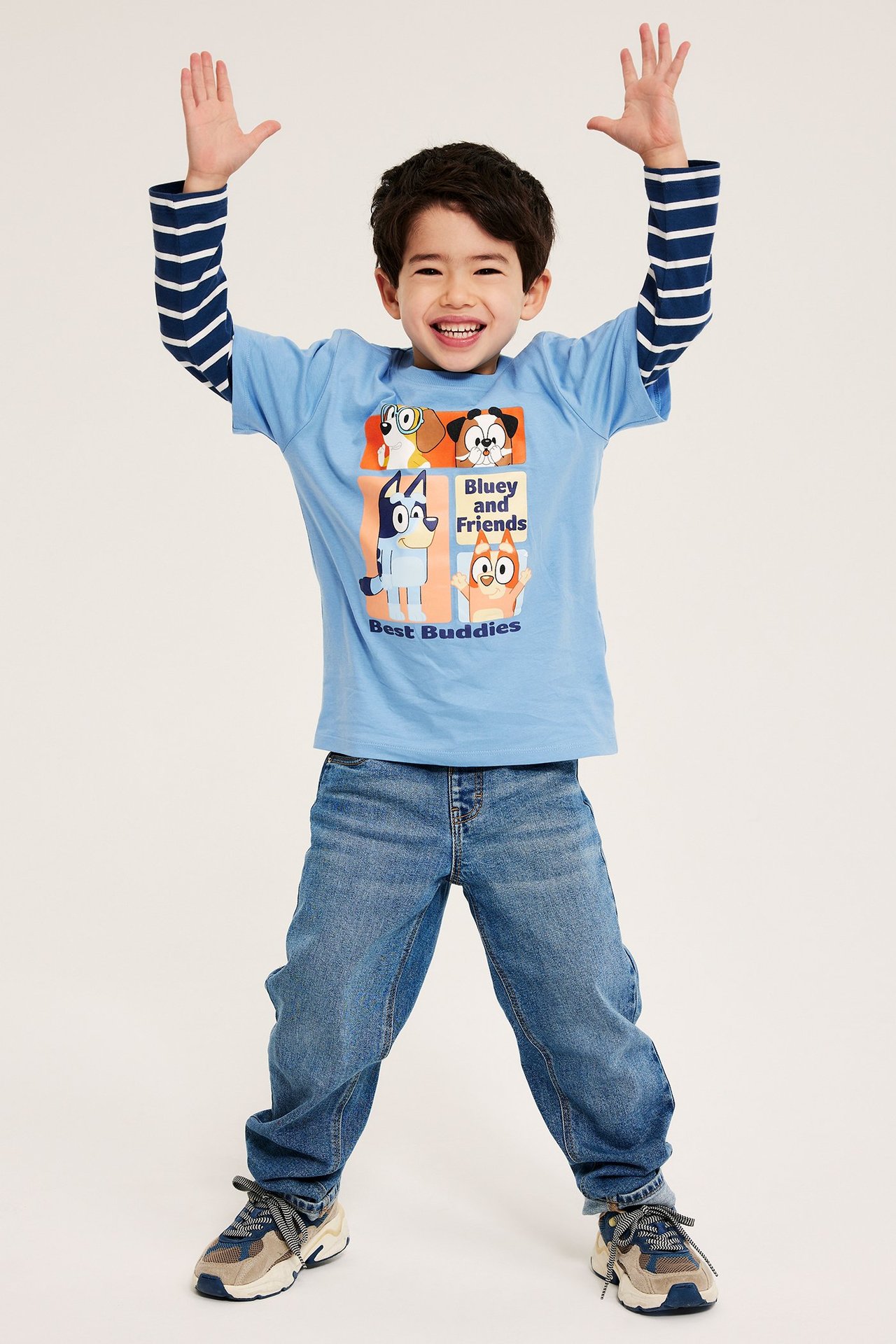 Widok z przodu dziecka w jasnoniebieskim T-shircie z nadrukiem Bluey, pasiastym longsleeve'ie, dżinsach i sneakersach.
