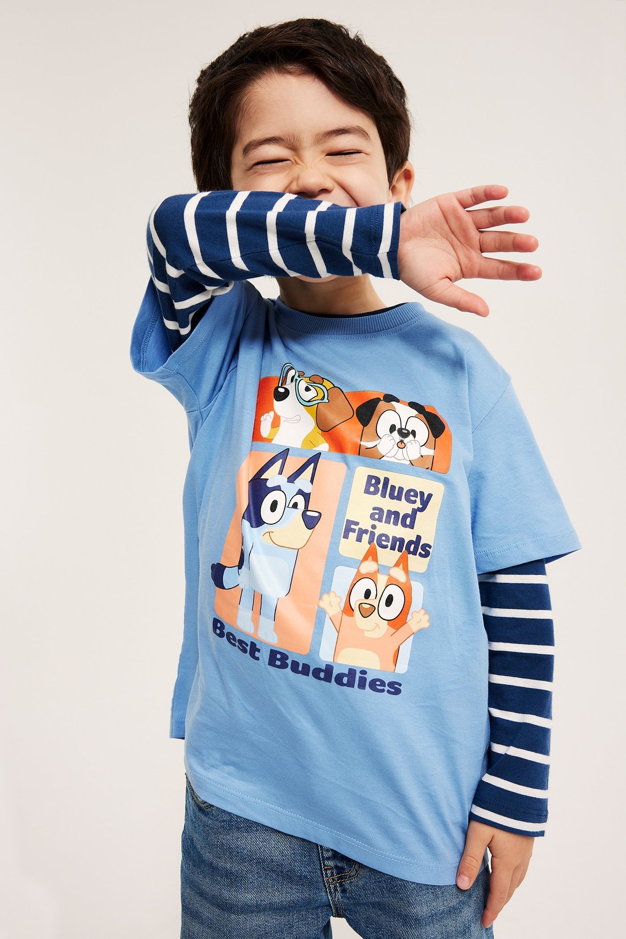 Widok z przodu: dziecko w jasnoniebieskiej koszulce z nadrukiem Bluey, na bluzce w paski i dżinsach.