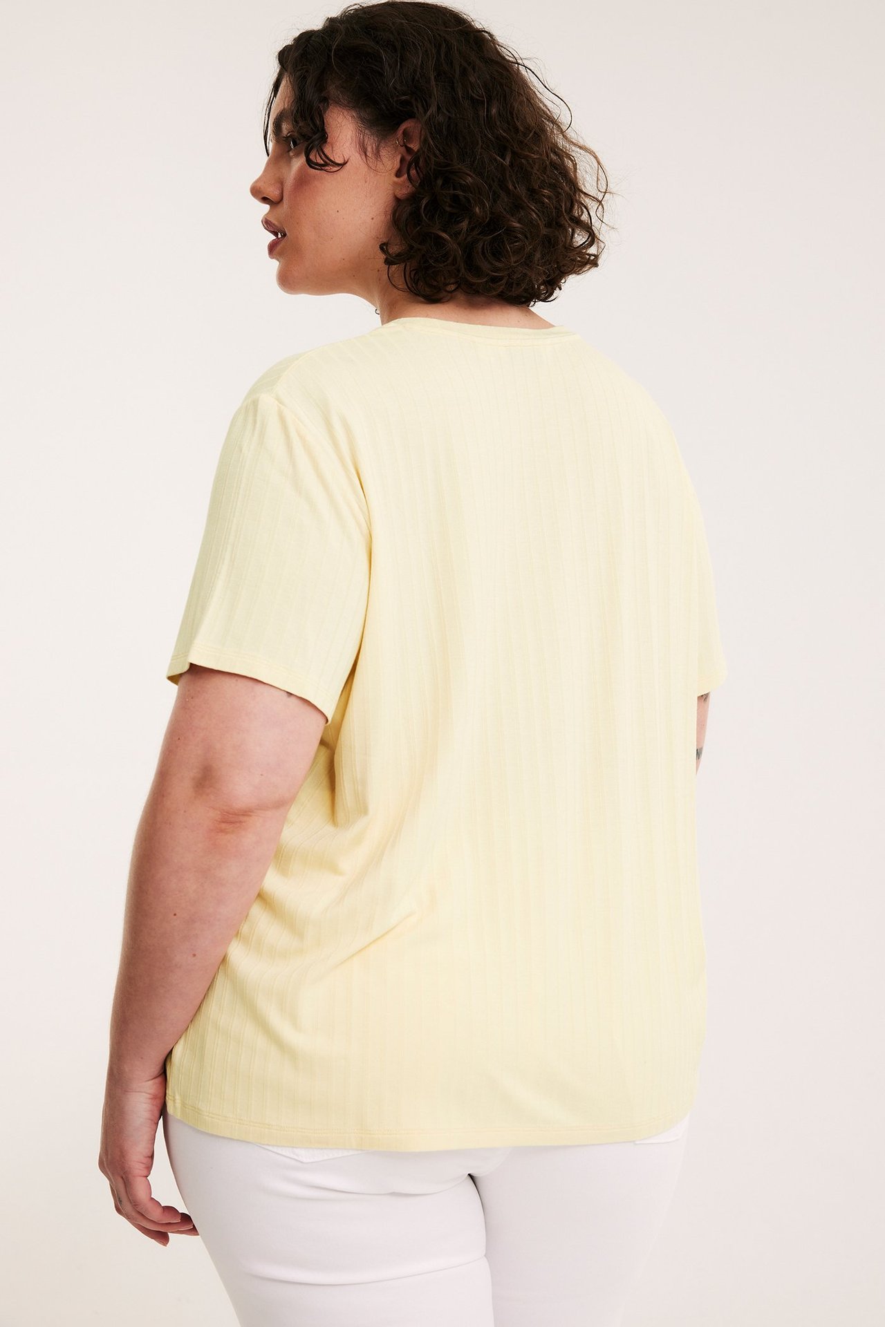 Sned bakvy av en kvinna i en ljusgul ribbad t-shirt och vita byxor.