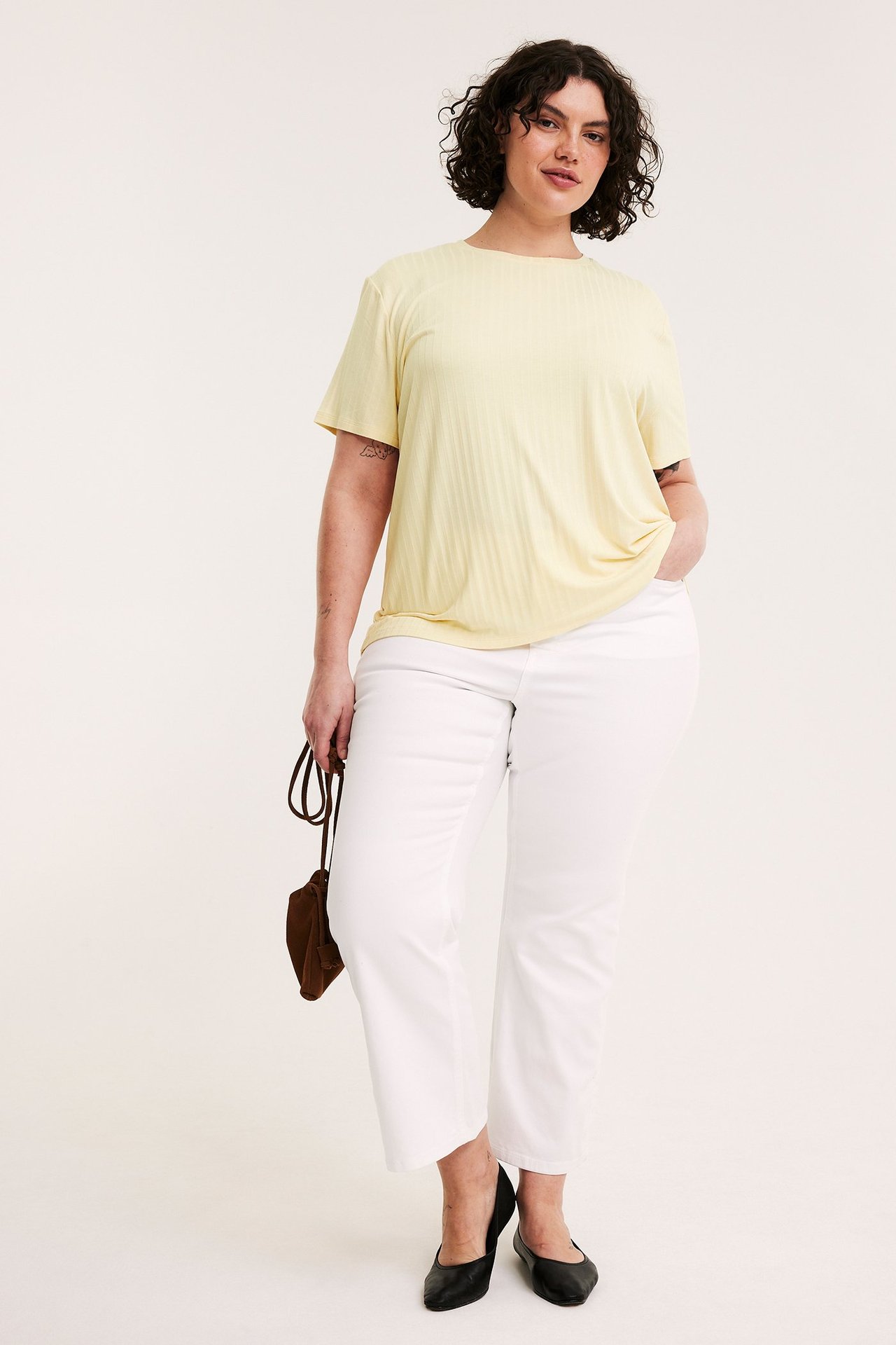 Framifrån: kvinna i ljusgul ribbad t-shirt, vita byxor, svarta skor och en brun väska.