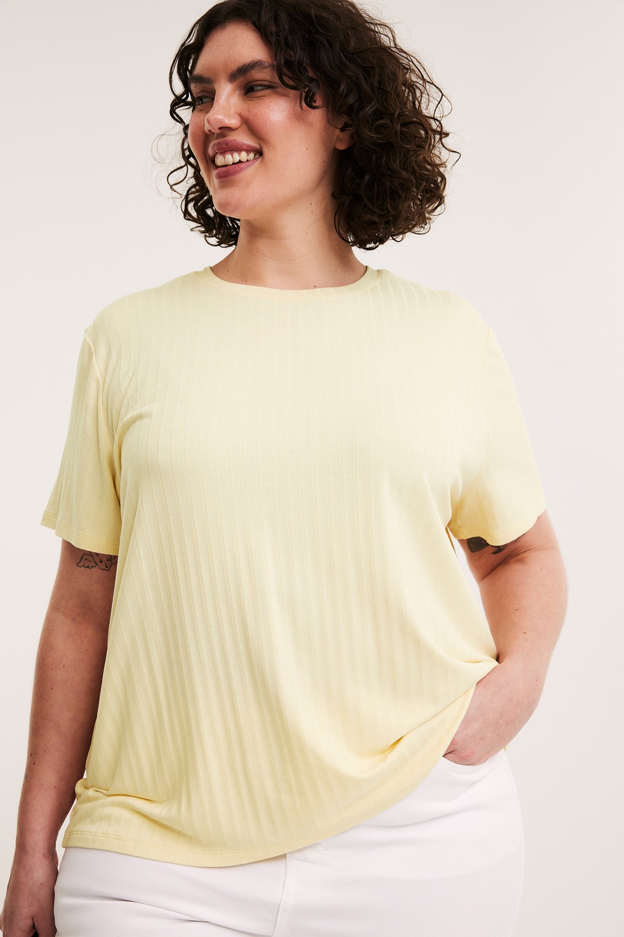 Framifrån, leende kvinna i en ljusgul ribbad t-shirt med boxig passform och vita byxor.