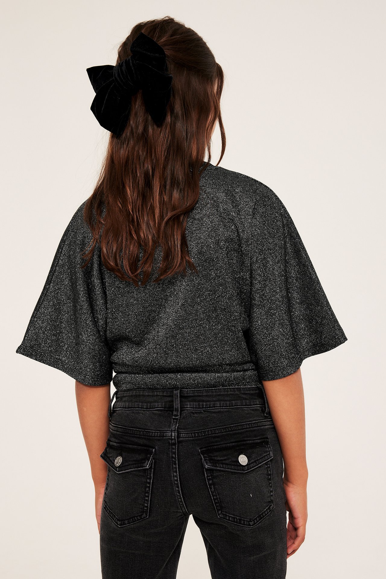 Ryggvy: Barn i svart glittrig topp med fjärilsärm, mörka jeans och svart sammetsrosett.