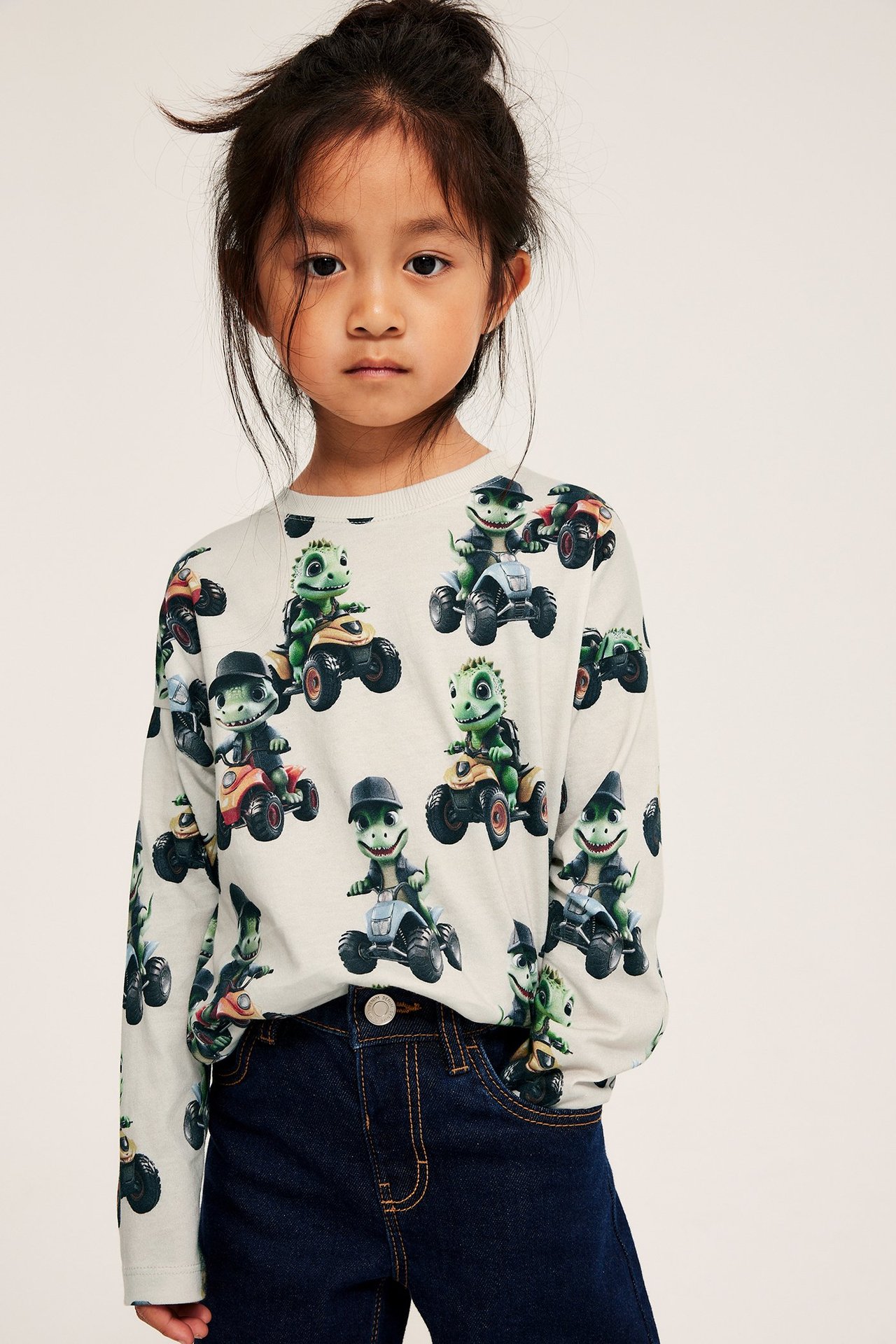 Frontbilde av et barn i en lys beige bomulls langermet t-skjorte med krokodille-ATV-print og mørkeblå jeans.