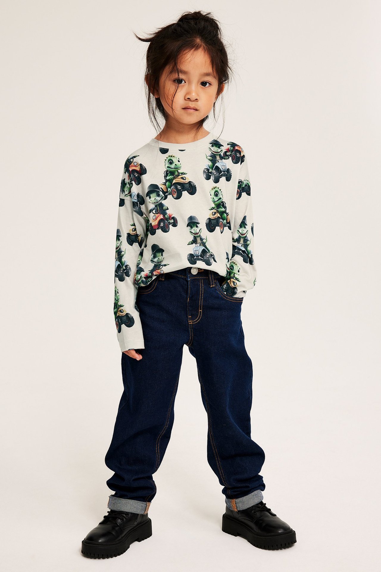 Frontvisning: Et barn i lys beige bomulls langermet t-skjorte med krokodillemønster, mørkeblå jeans og svarte sko.