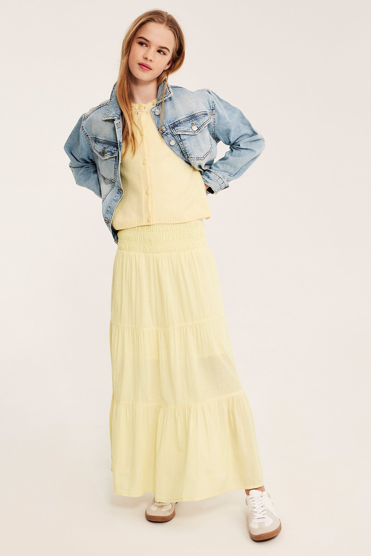 Lapsi yllään vaaleankeltainen röyhelöinen maksihame rypytetyllä vyötäröllä, keltainen neuletakki ja vaaleansininen farkkutakki.