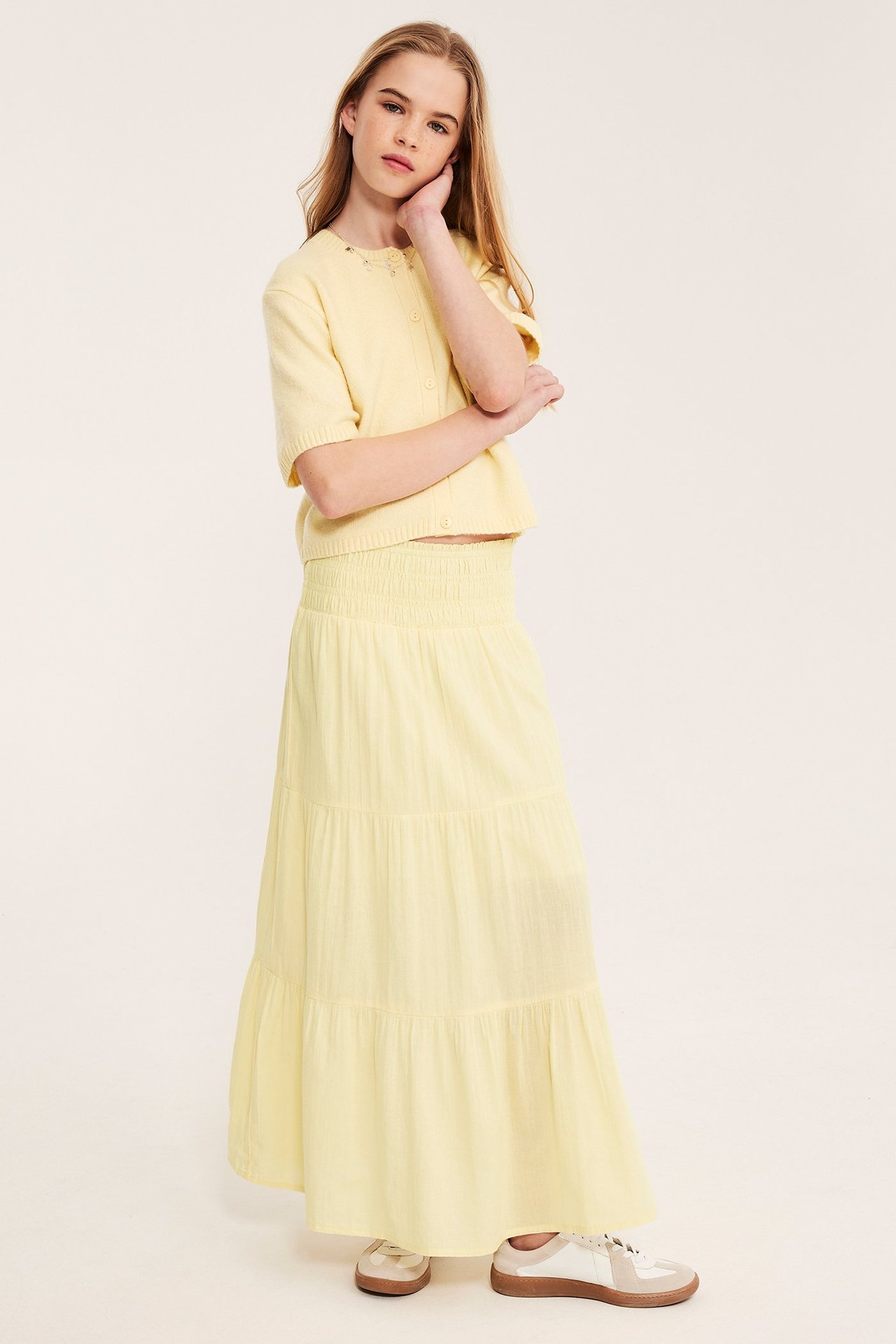 Widok z przodu: Dziecko w jasnożółtej bawełnianej warstwowej spódnicy maxi, pasującym krótkim kardiganie i białych i beżowych sneakersach.