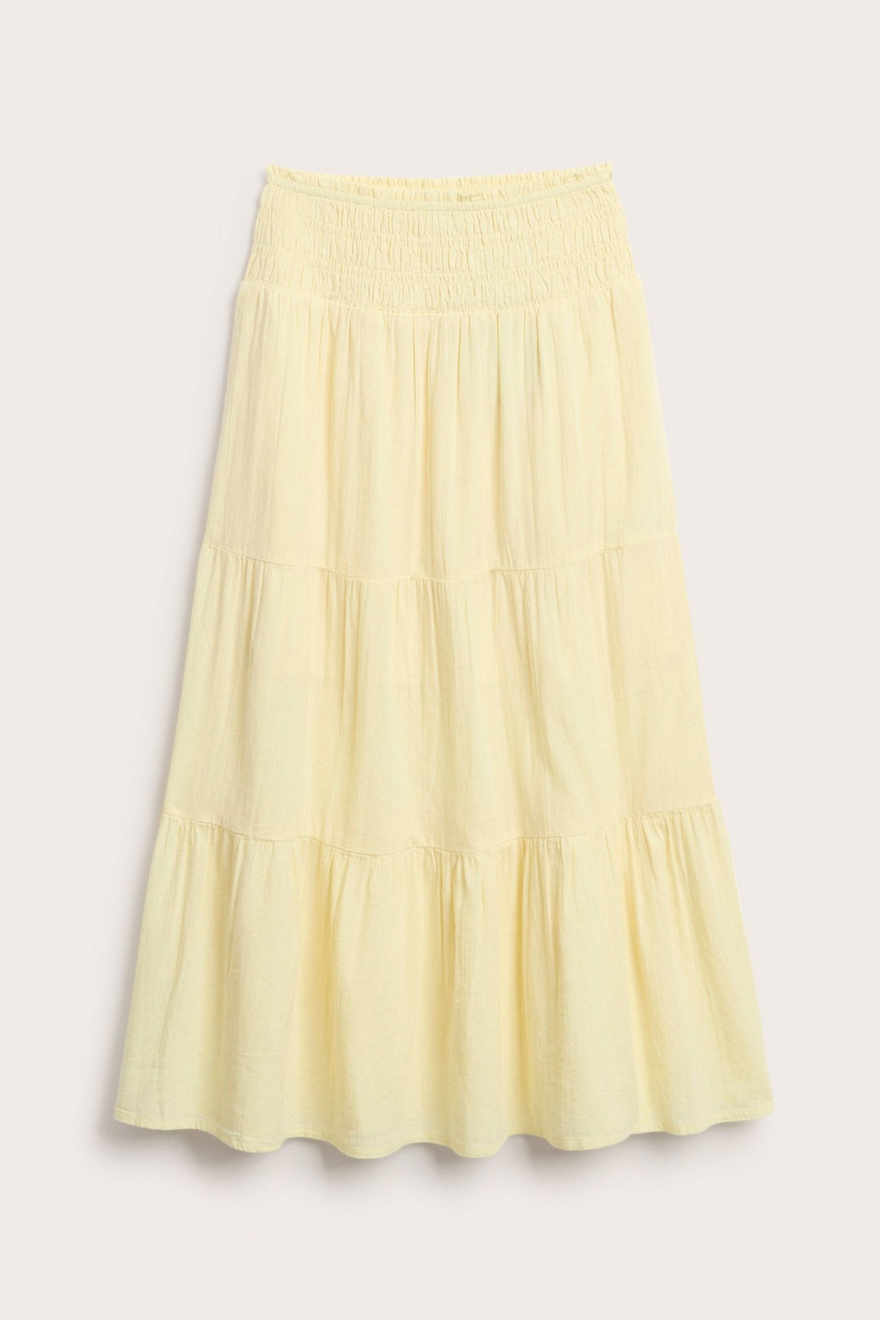 Frontvy av en ljusgul maxikjol i vävd bomull med smockad midja och volanger.