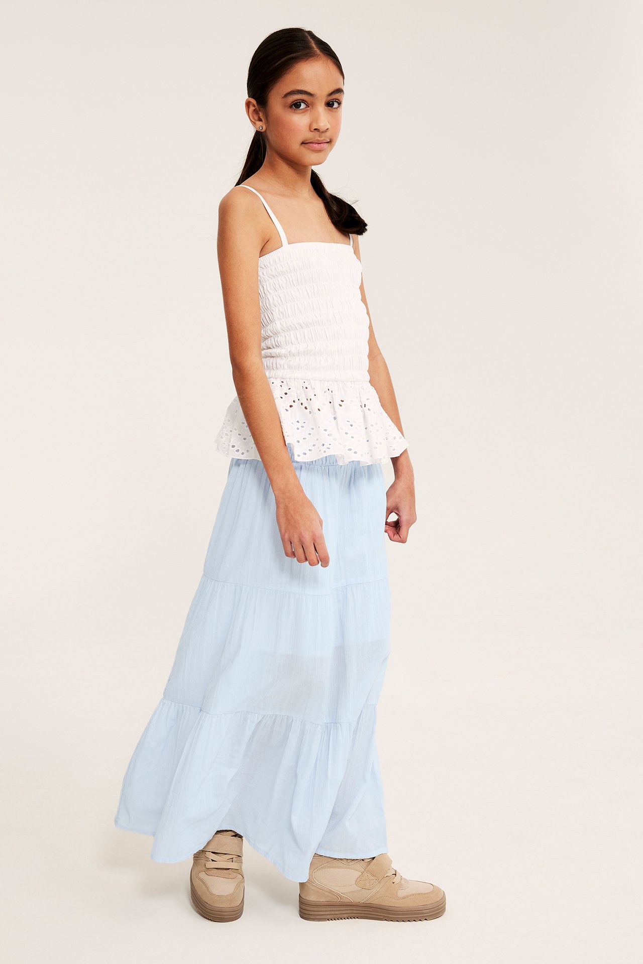 Kid i ljusblå volangmaxikjol med smockad midja, vit smockad topp och beiga sneakers.