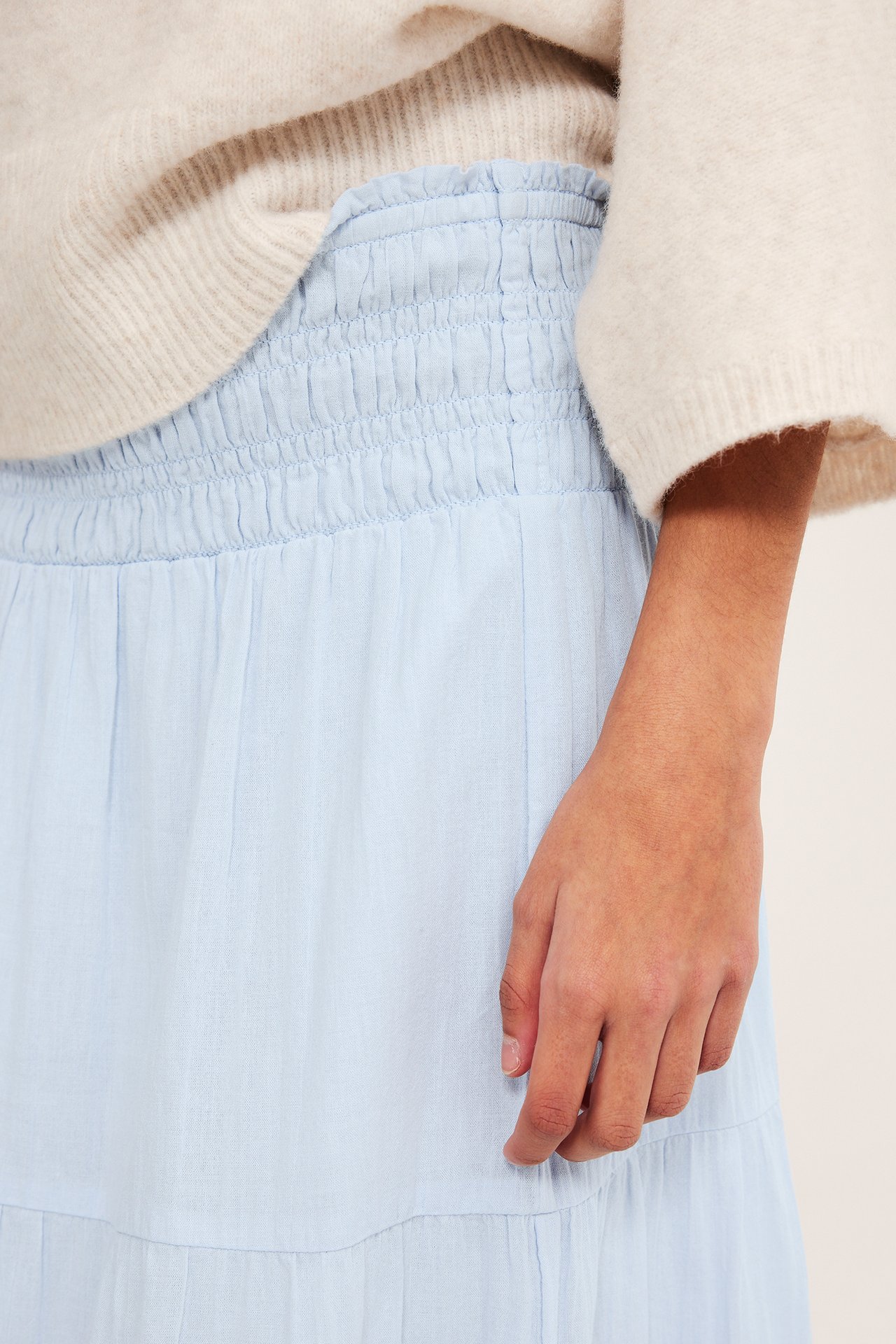 Zbliżenie kobiety w jasnoniebieskiej marszczonej spódnicy maxi z falbankami i kremowym swetrze.