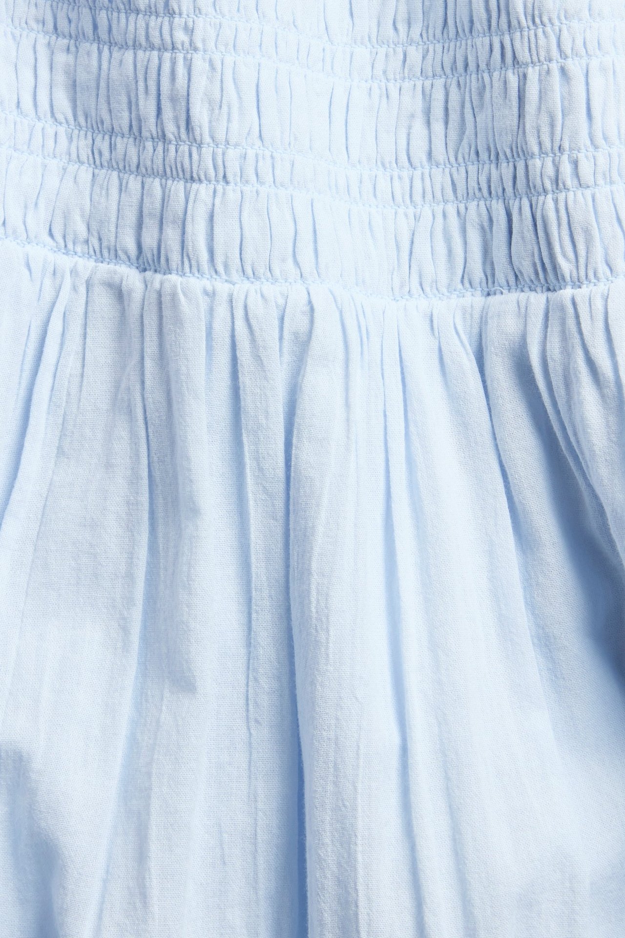 Närbild av en ljusblå maxikjol med smockad midja och veckat vävt bomullstyg.