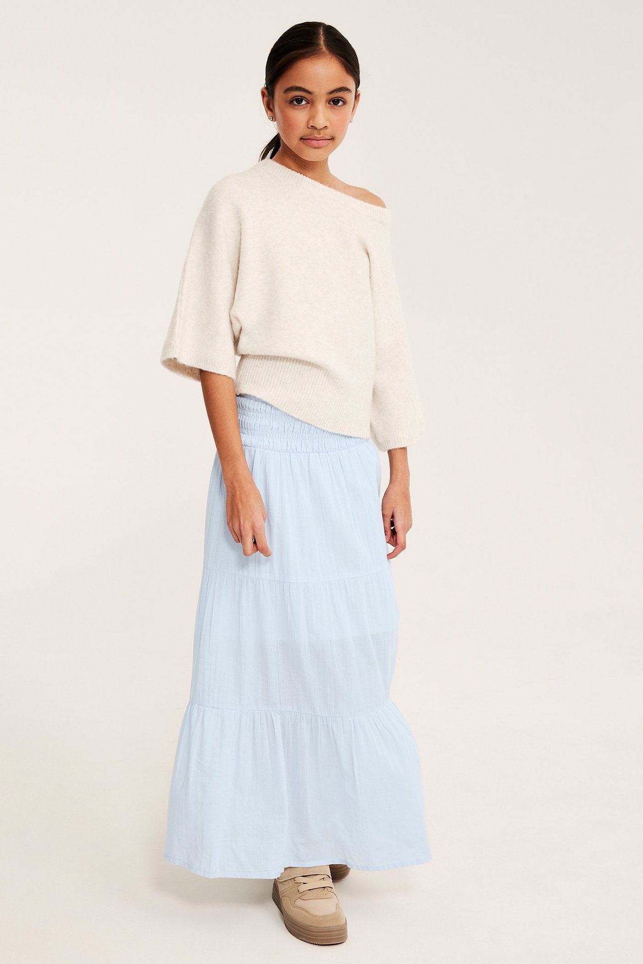 Framifrån: Barn i ljusblå volangmaxikjol med smockad midja, offwhite tröja och beiga sneakers.