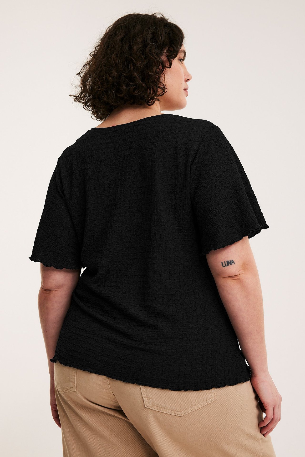 Bakifrån på kvinna i svart strukturerad kortärmad topp med vågiga kanter och beiga byxor.