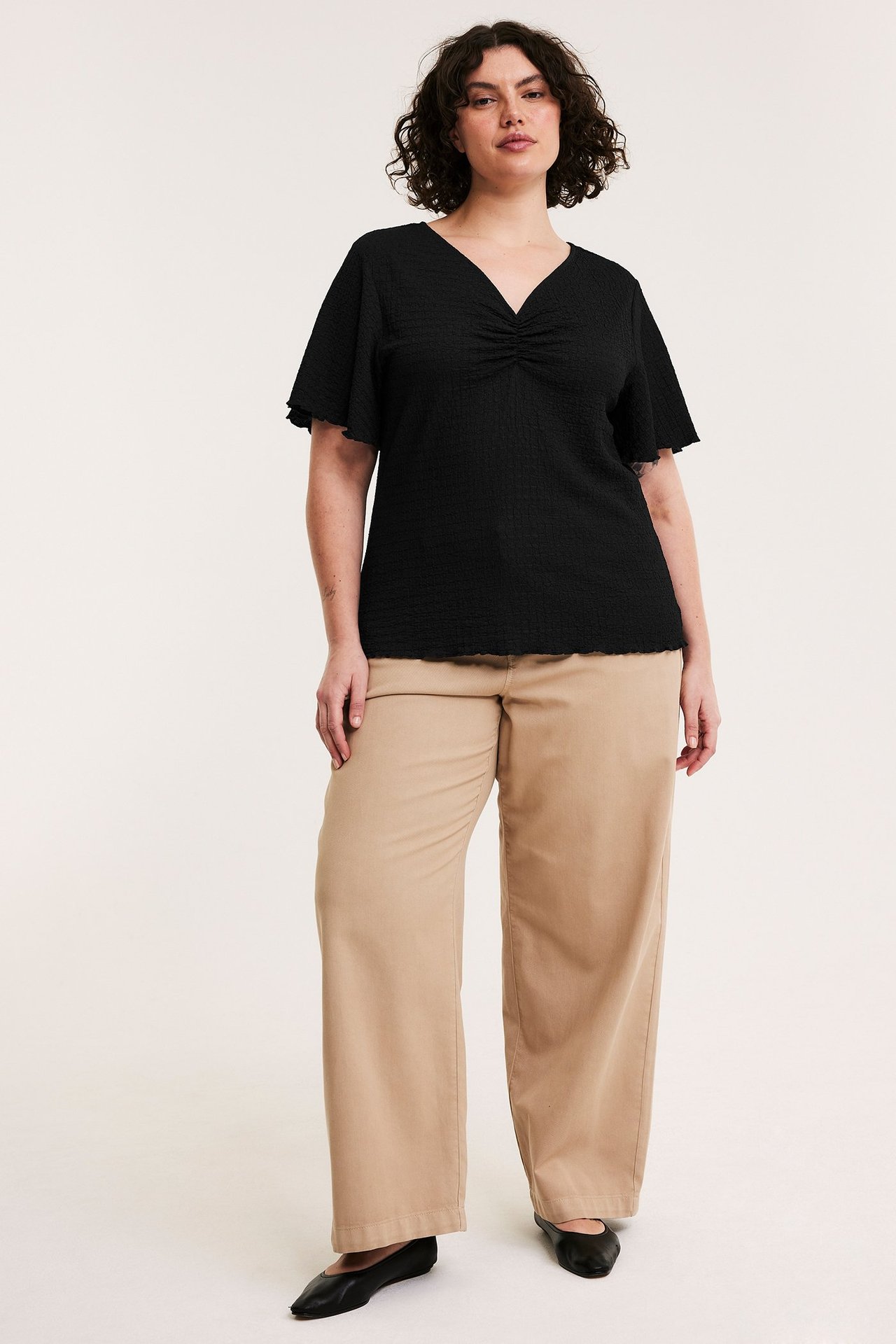 Framifrån: Kvinna i svart strukturerad kortärmad topp, beiga vida byxor och svarta ballerinaskor.
