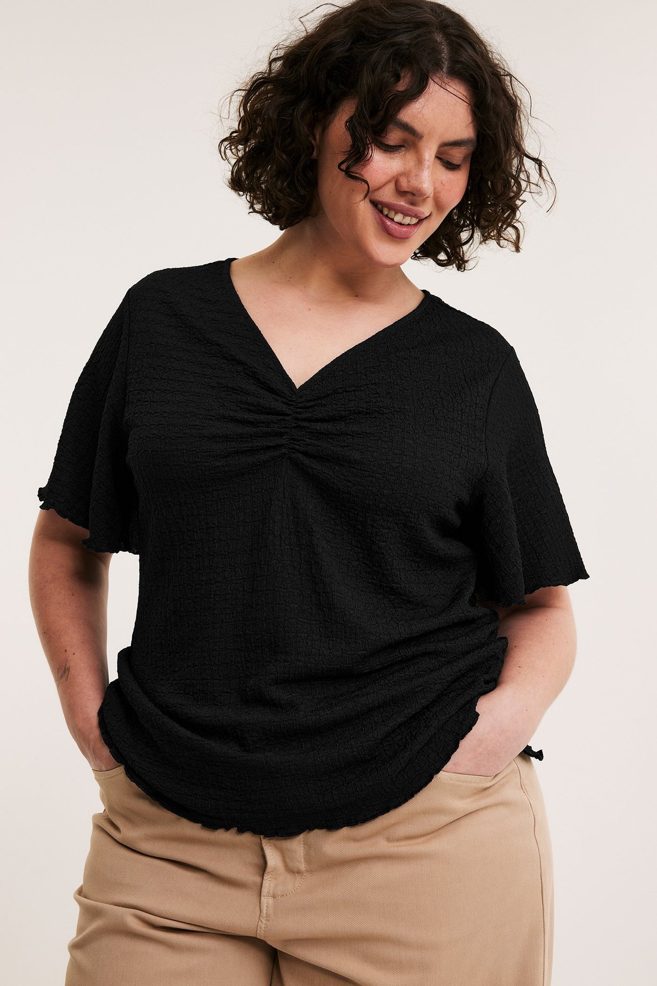Czarny teksturowany top z dekoltem w serek, rozszerzanymi rękawami, marszczony przód. Beżowe spodnie.