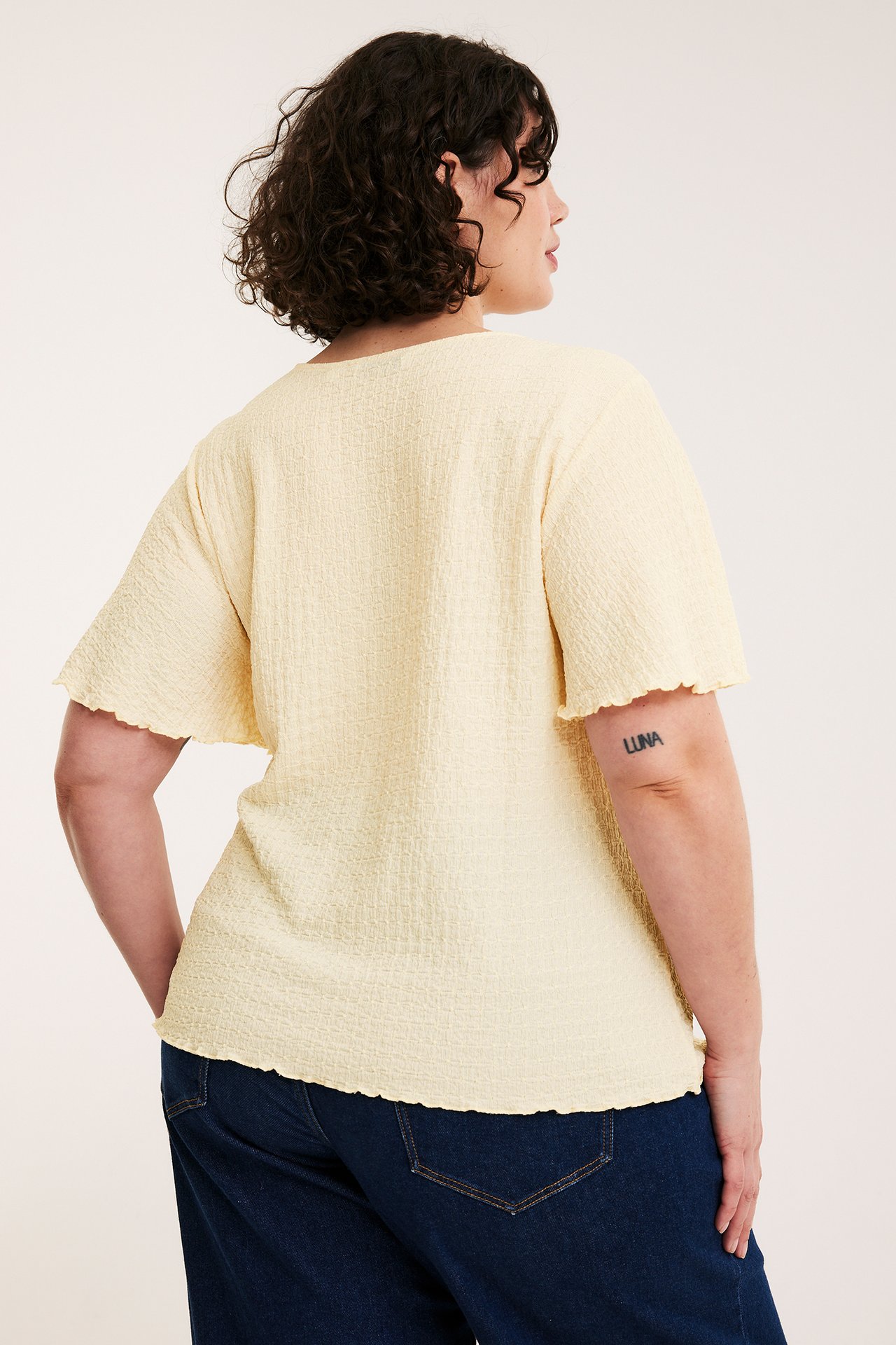 Ryggvy av kvinna i ljusgul strukturerad kortärmad topp med vågiga kanter och mörkblå jeans.
