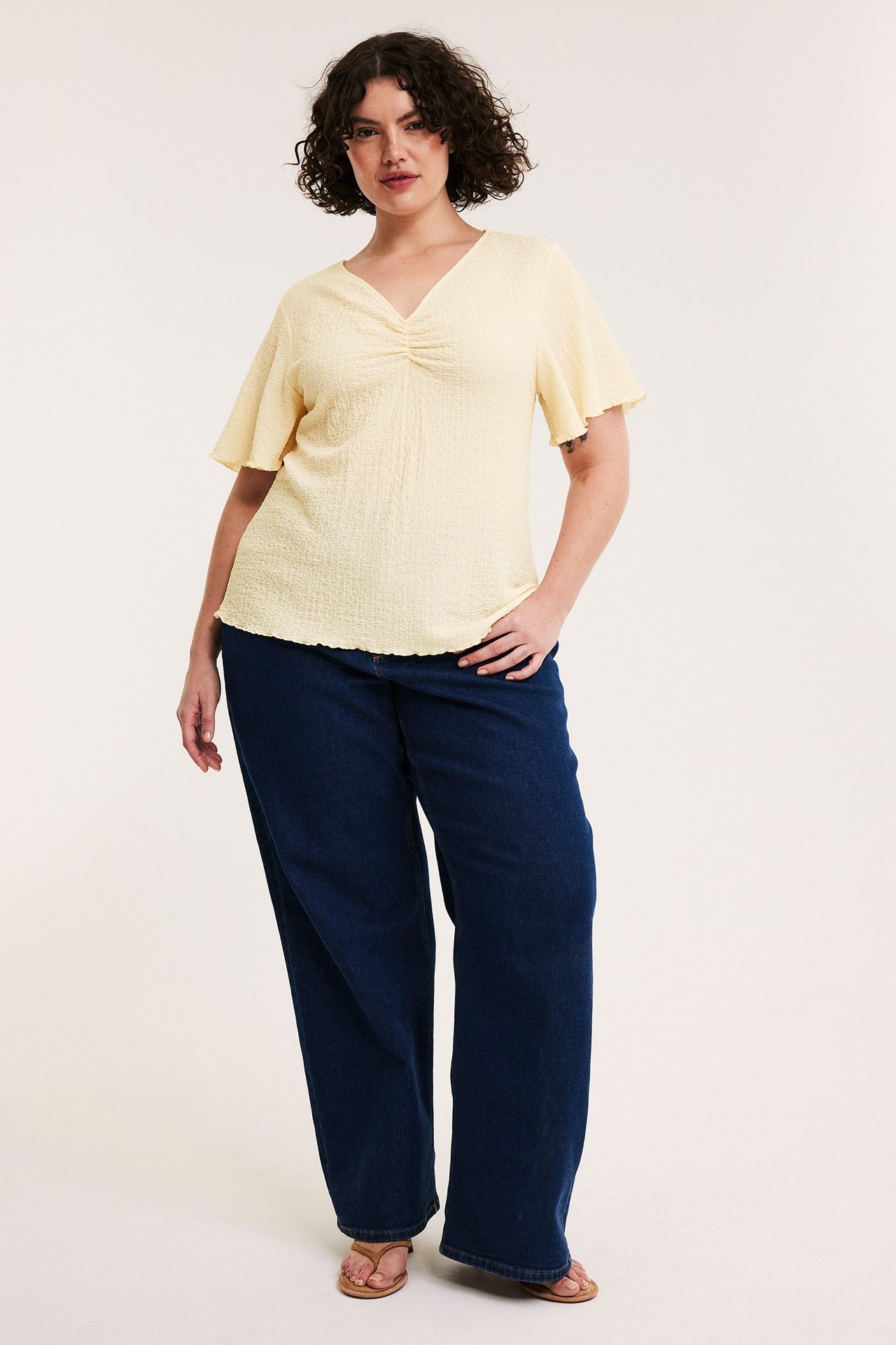 Edestäpäin: nainen yllään vaaleankeltainen kuvioitu toppi, tummansiniset farkut ja ruskeat sandaalit.