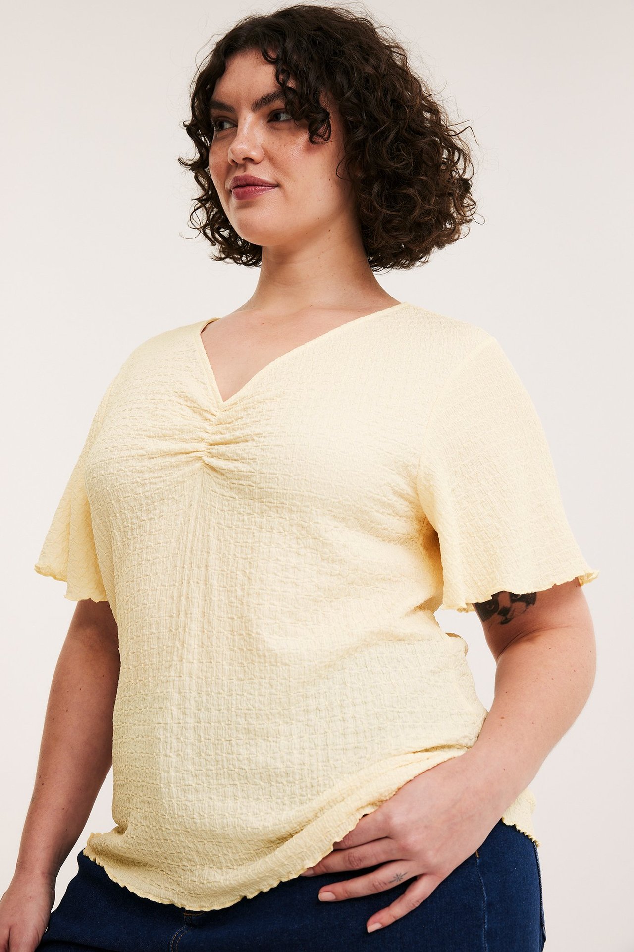 Kobieta nosi jasnożółty, krótki top z marszczonym dekoltem V i ciemnoniebieskie jeansy.