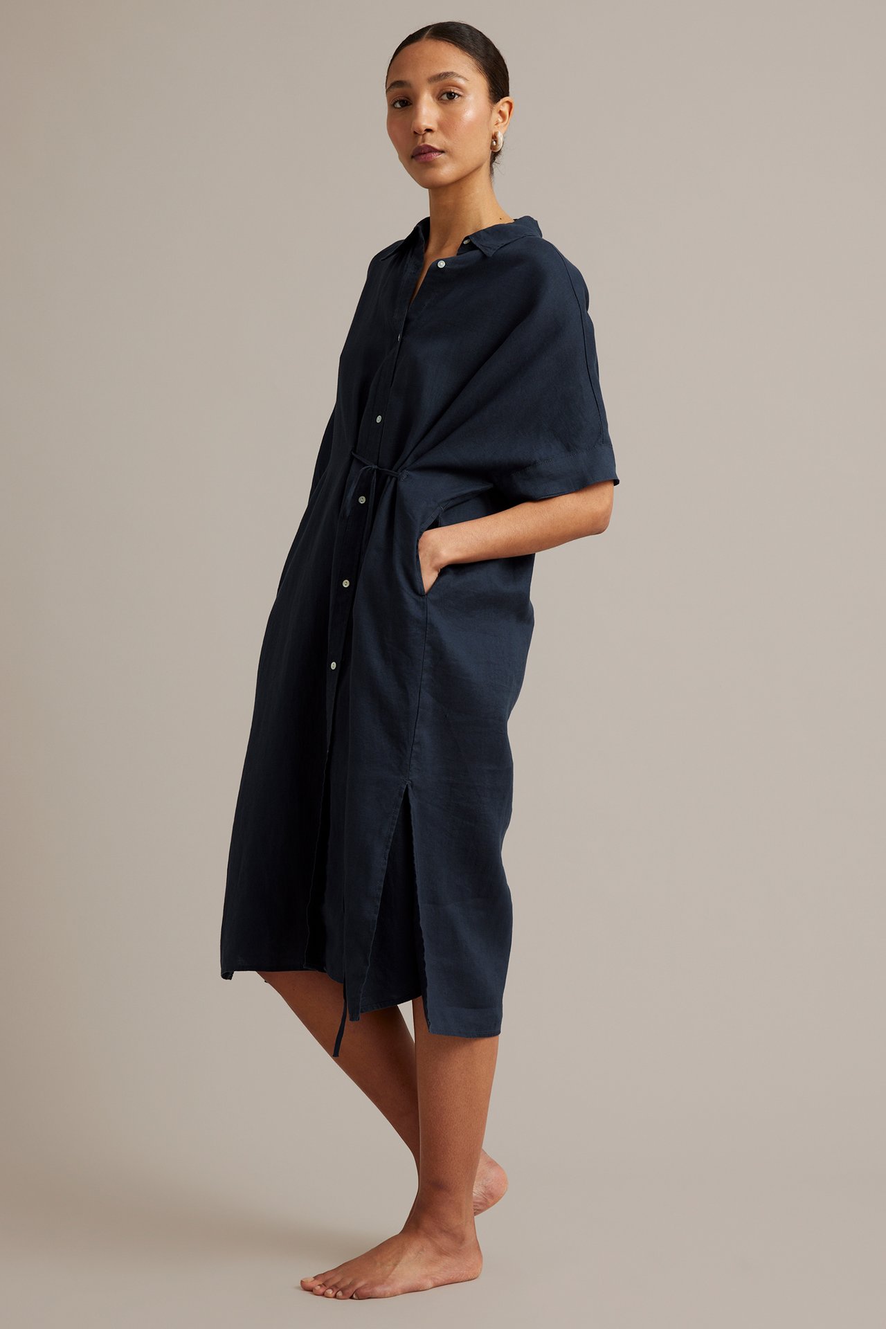 Sidovy: kvinna i mörkblå linneskjortklänning med korta ärmar, knytdragsko i midjan och sidoslitsar.