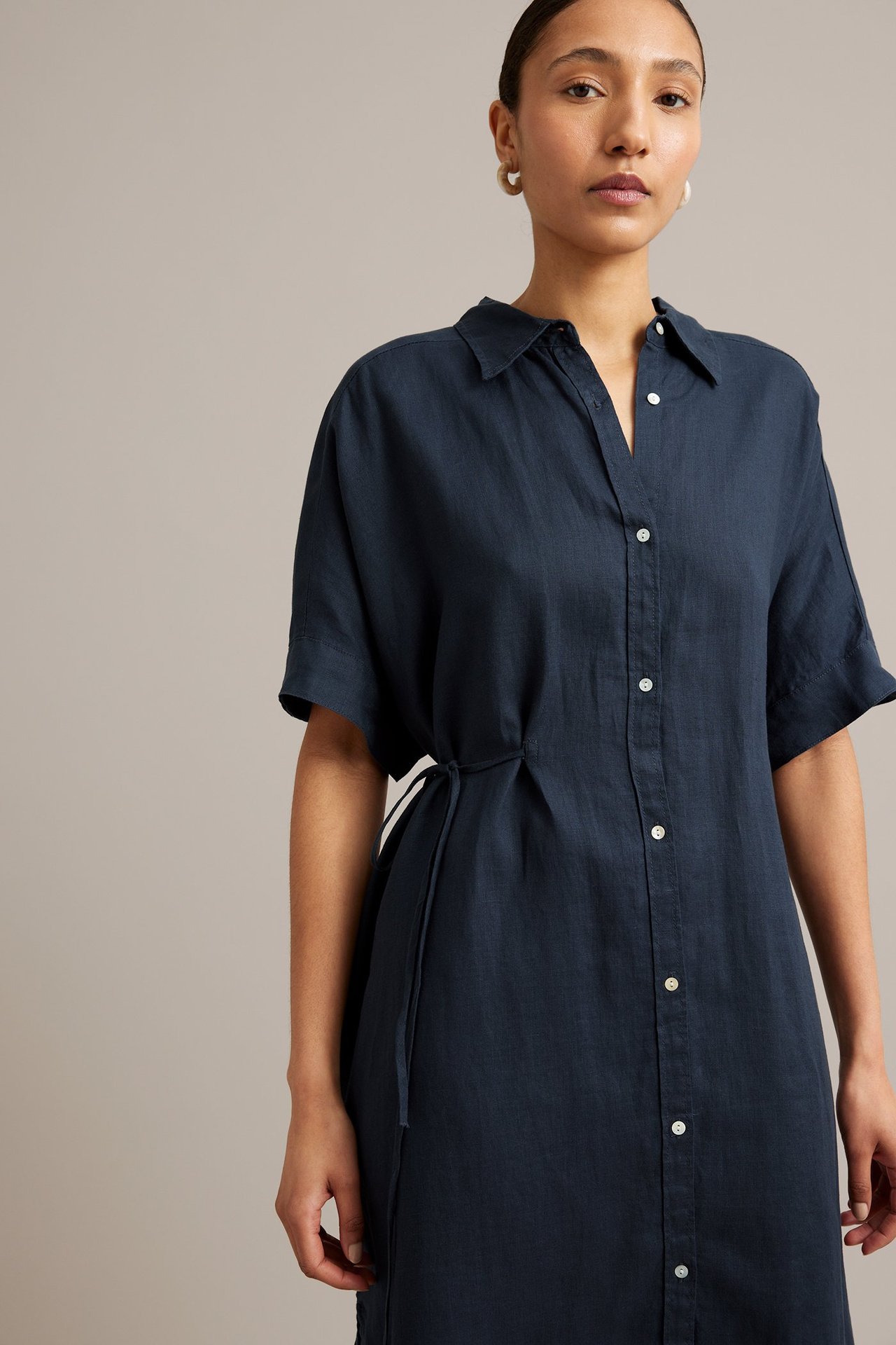 Kvinna i mörkblå linneskjortklänning med korta ärmar, vita knappar, knuten midja och ringörhängen.